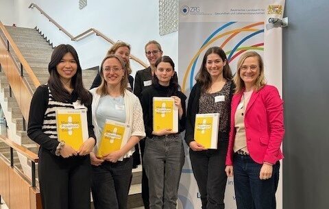 Sieben Personen stehen in einem Innenraum vor einer Treppe. Vorne stehen vier junge Frauen, Absolventinnen des Masters Gesundheitsförderung, mit gelben Zertifikaten in der Hand und lächeln. Drei Erwachsene stehen hinter ihnen. Rechts ist ein Banner der Präventionskonferenz Bayern mit bunten Ringen zu sehen. at Hochschule Coburg