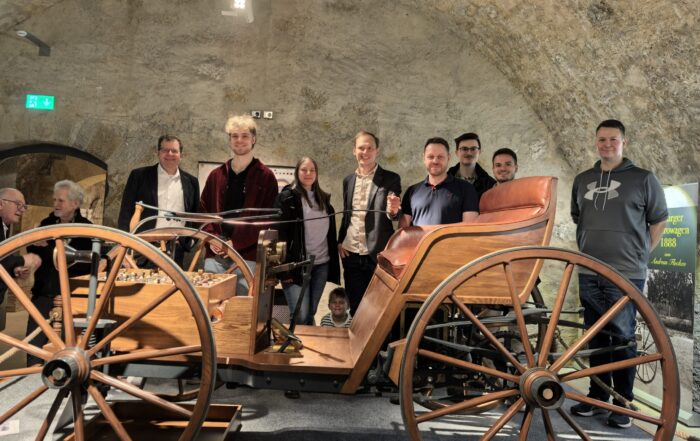 Eine Gruppe von neun Personen steht lächelnd um ein altmodisches hölzernes Auto, das unter einem steinernen Torbogen ausgestellt ist, als ob es aus alten Fotos in einer museumsähnlichen Umgebung stammt. at Hochschule Coburg