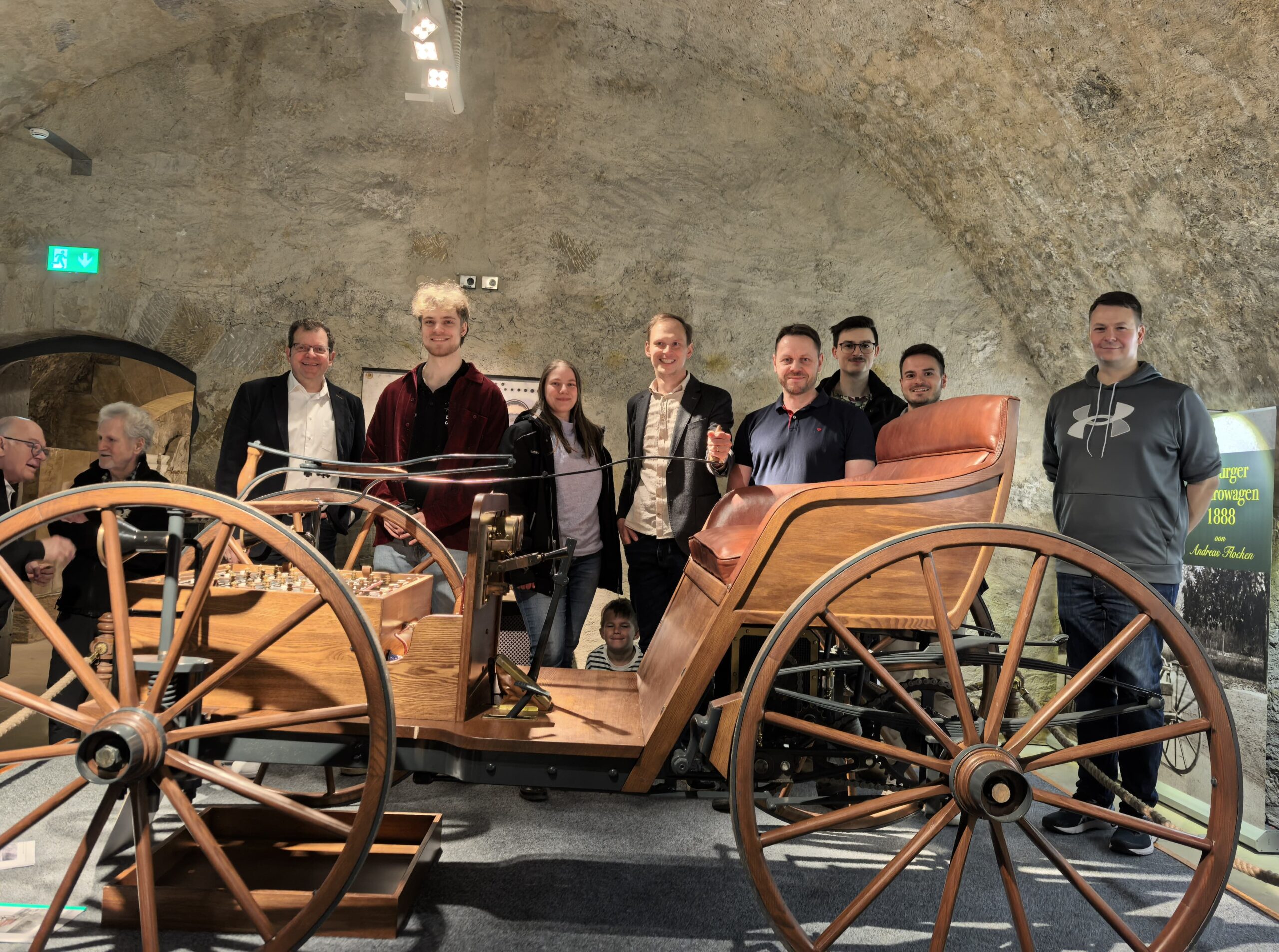Eine Gruppe von neun Personen steht lächelnd um ein altmodisches hölzernes Auto, das unter einem steinernen Torbogen ausgestellt ist, als ob es aus alten Fotos in einer museumsähnlichen Umgebung stammt. at Hochschule Coburg