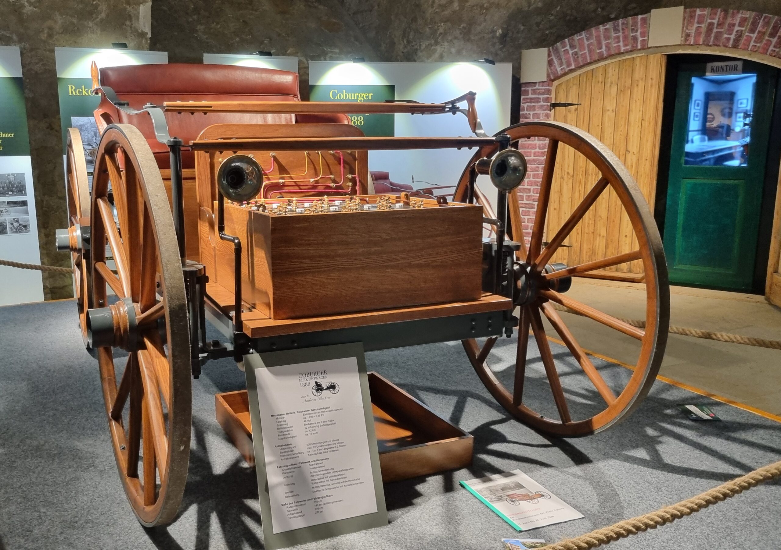Eine alte Holzkutsche mit großen Speichenrädern wird in einer Museumsumgebung ausgestellt, ihre Rekonstruktion basiert auf alten Fotos. Informationsschilder und Seile umgeben das Exponat, mit einer grünen Tür und einem gewölbten Backsteineingang im Hintergrund. at Hochschule Coburg