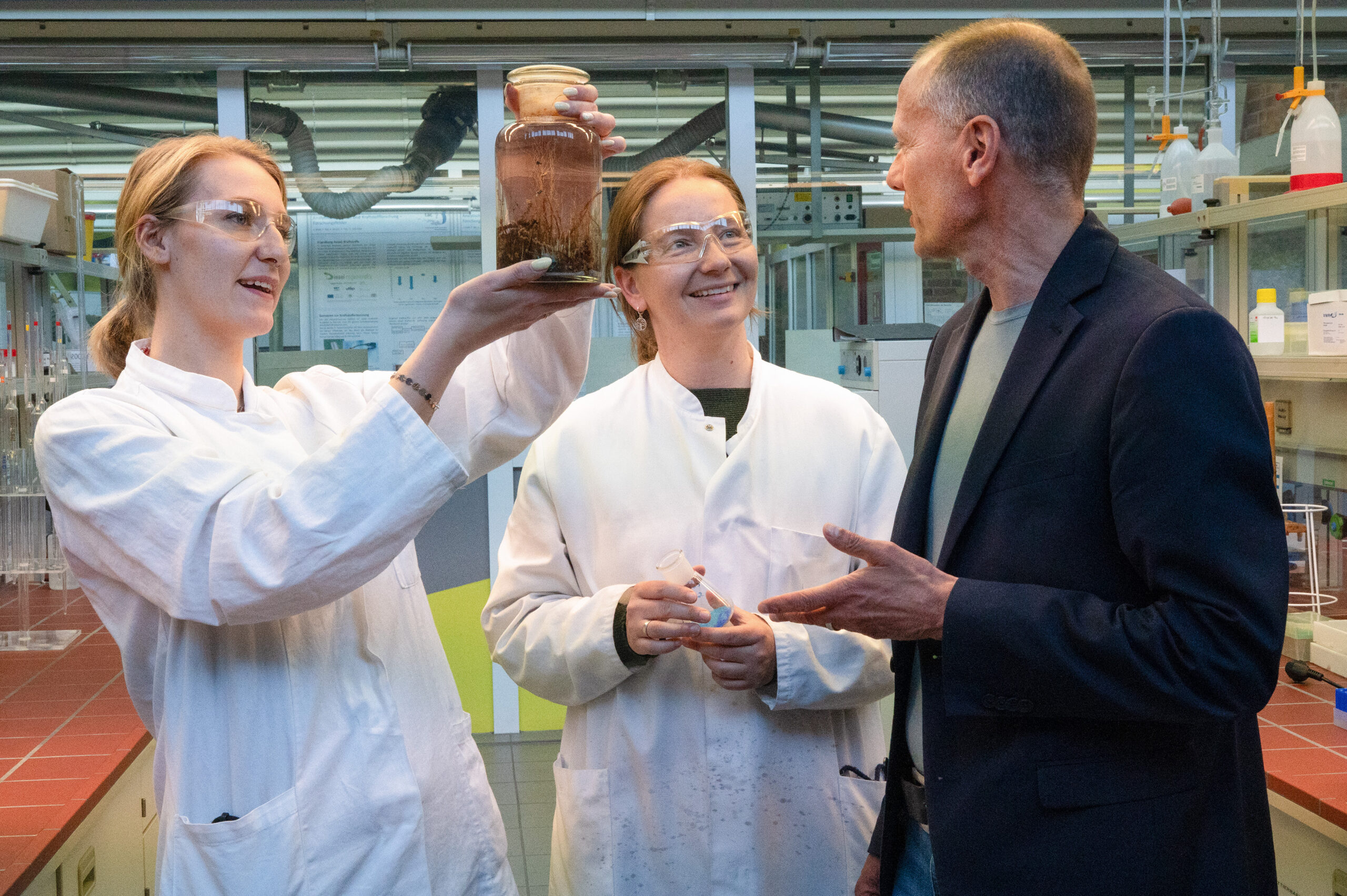 Während der Langen Nacht der Wissenschaften in Coburg untersuchen zwei Wissenschaftler in Laborkitteln und mit Schutzbrillen ein Glasgefäß mit Erde und Wurzeln, während ein Mann im Blazer in einem mit wissenschaftlichen Geräten ausgestatteten Labor über ihre Ergebnisse diskutiert. at Hochschule Coburg
