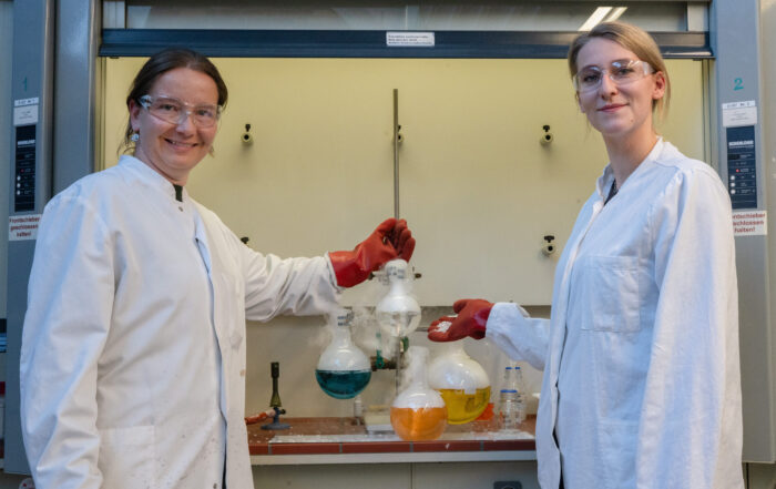 Zwei Personen in Laborkitteln und mit Schutzbrillen stehen in einem Coburger Labor und halten lächelnd Glaskolben mit blauen, gelben und klaren Flüssigkeiten in der Hand - und fangen damit den Forschergeist der Langen Nacht der Wissenschaften ein. Im Hintergrund sind Laborgeräte zu sehen. at Hochschule Coburg