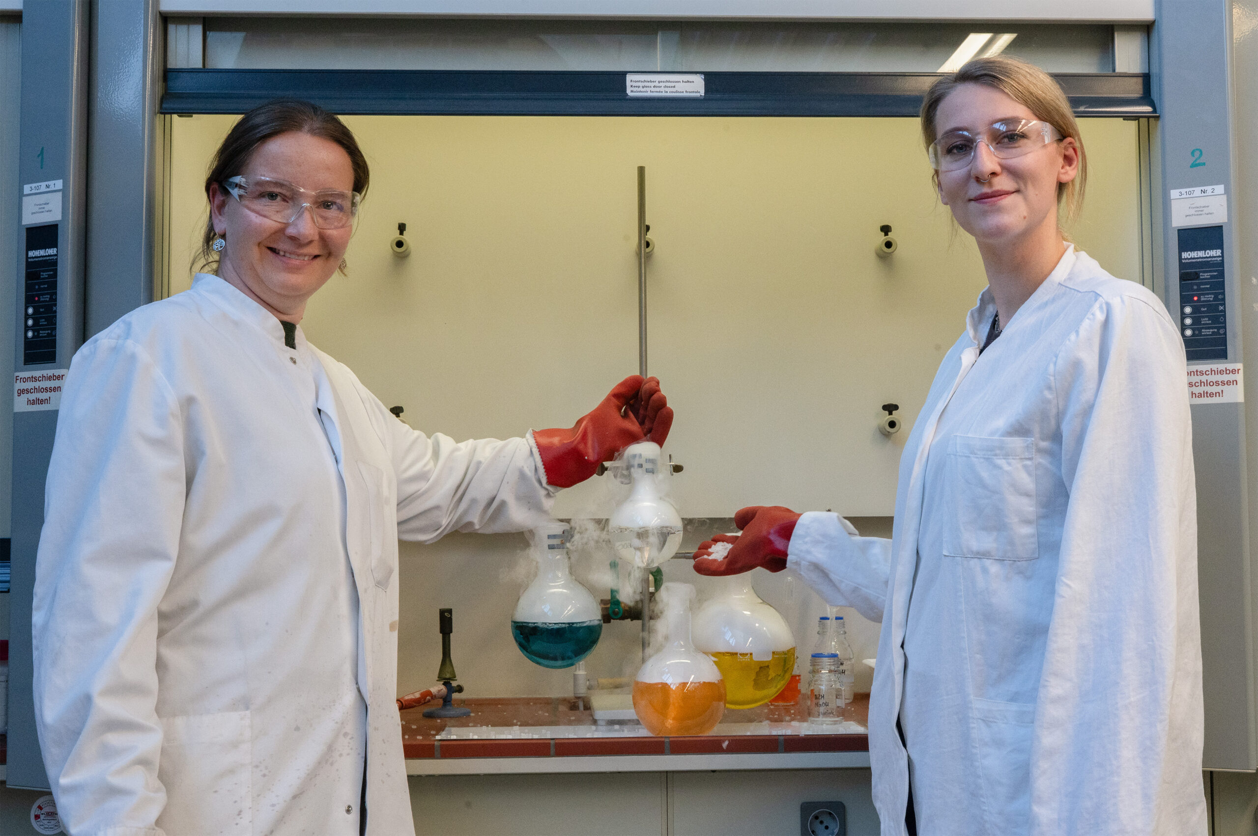 Zwei Personen in Laborkitteln und mit Schutzbrillen stehen in einem Coburger Labor und halten lächelnd Glaskolben mit blauen, gelben und klaren Flüssigkeiten in der Hand - und fangen damit den Forschergeist der Langen Nacht der Wissenschaften ein. Im Hintergrund sind Laborgeräte zu sehen. at Hochschule Coburg