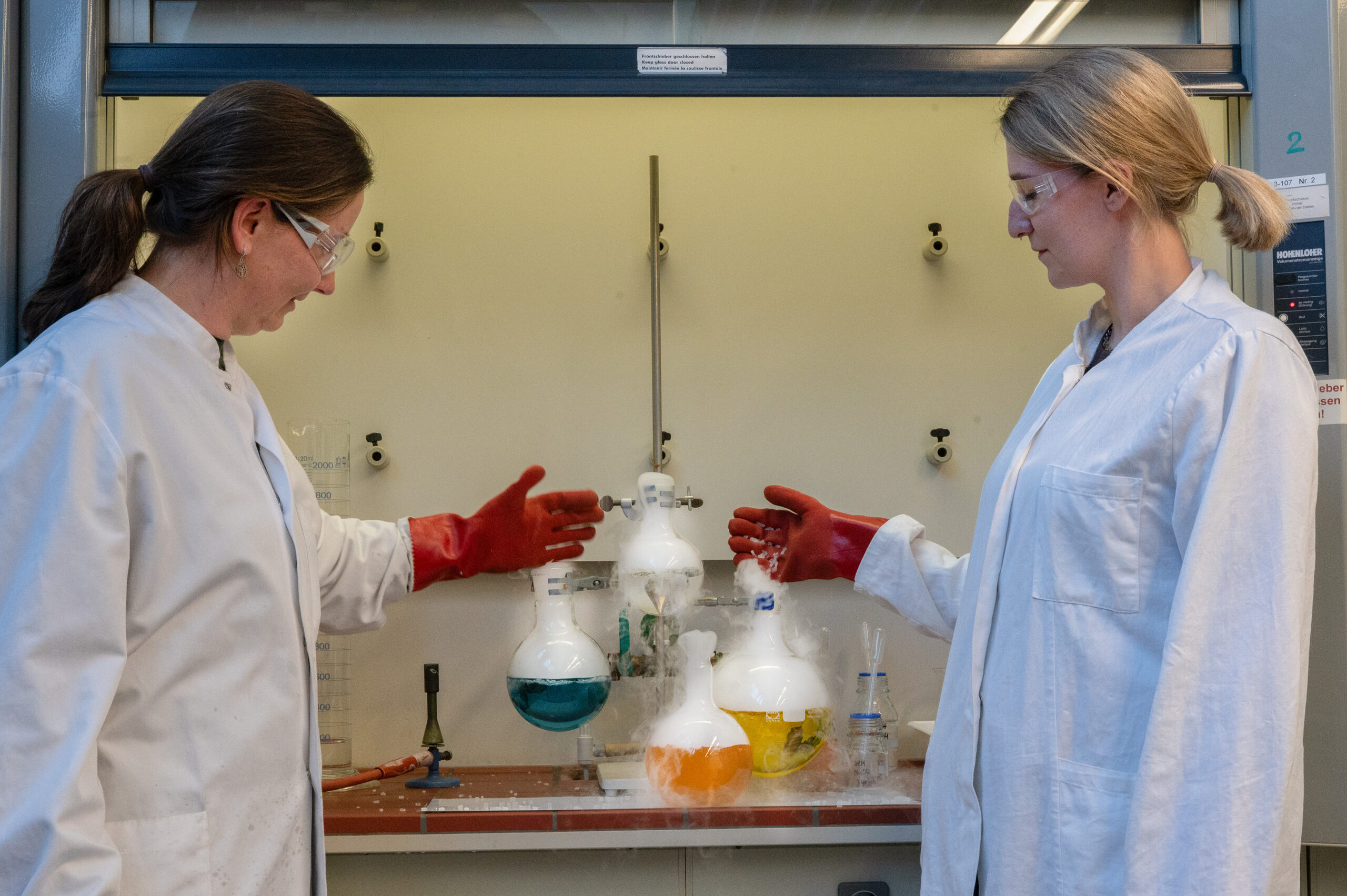 Während der Langen Nacht der Wissenschaften in Coburg stehen zwei Personen in Laborkitteln und mit Schutzbrillen vor einem Abzug, gestikulieren in Richtung vibrierender Kolben, aus denen Dämpfe austreten, und tragen rote Schutzhandschuhe. at Hochschule Coburg