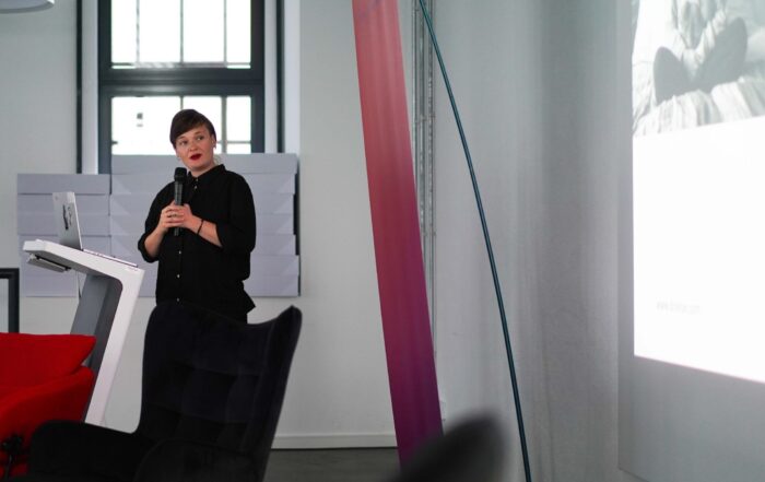 Eine Frau steht an einem Podium mit einem Mikrofon und hält einen Vortrag über Endometriose vor einem Publikum in einem modernen, hellen Raum mit roten und schwarzen Stühlen und einem projizierten Bild an einer weißen Wand. at Hochschule Coburg