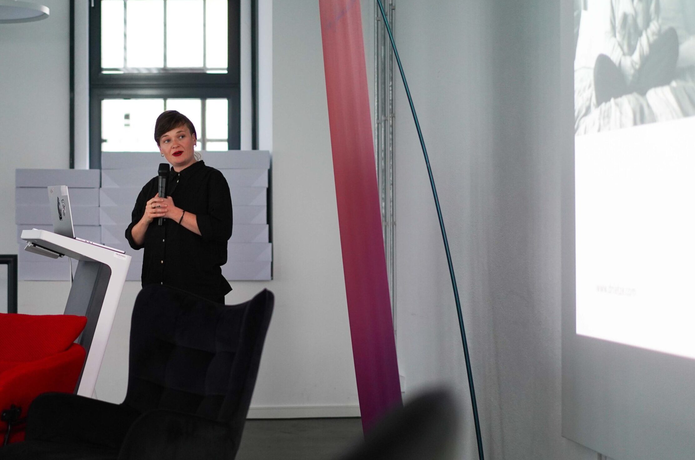 Eine Frau steht an einem Podium mit einem Mikrofon und hält einen Vortrag über Endometriose vor einem Publikum in einem modernen, hellen Raum mit roten und schwarzen Stühlen und einem projizierten Bild an einer weißen Wand. at Hochschule Coburg