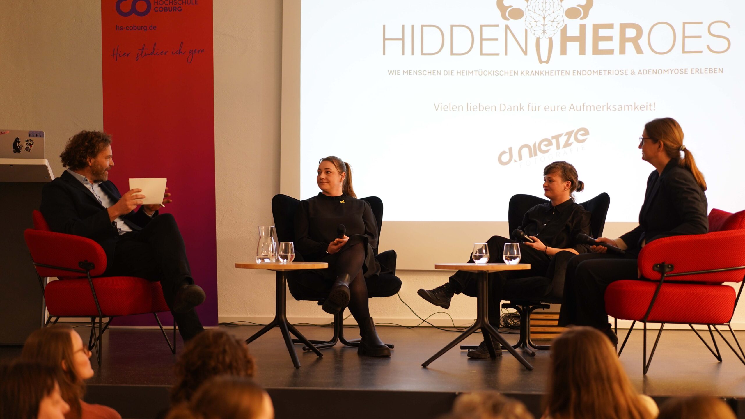 Vier Personen sitzen auf der Bühne auf Stühlen und führen eine Podiumsdiskussion über Endometriose und Adenomyose. Hinter ihnen zeigt eine große Leinwand das Logo und den Text von "Hidden Heroes". Im Vordergrund ist ein aufmerksames Publikum zu sehen. at Hochschule Coburg
