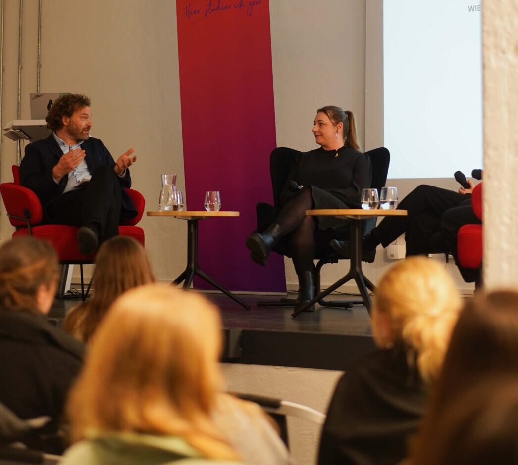 Zwei Personen sitzen auf roten Stühlen auf einer Bühne und führen eine Podiumsdiskussion über Adenomyose und Endometriose. Das Publikum beobachtet, wie hinter den Rednern in diesem modernen Veranstaltungsraum ein Bildschirm und ein rosa Banner mit Text eingeblendet werden. at Hochschule Coburg