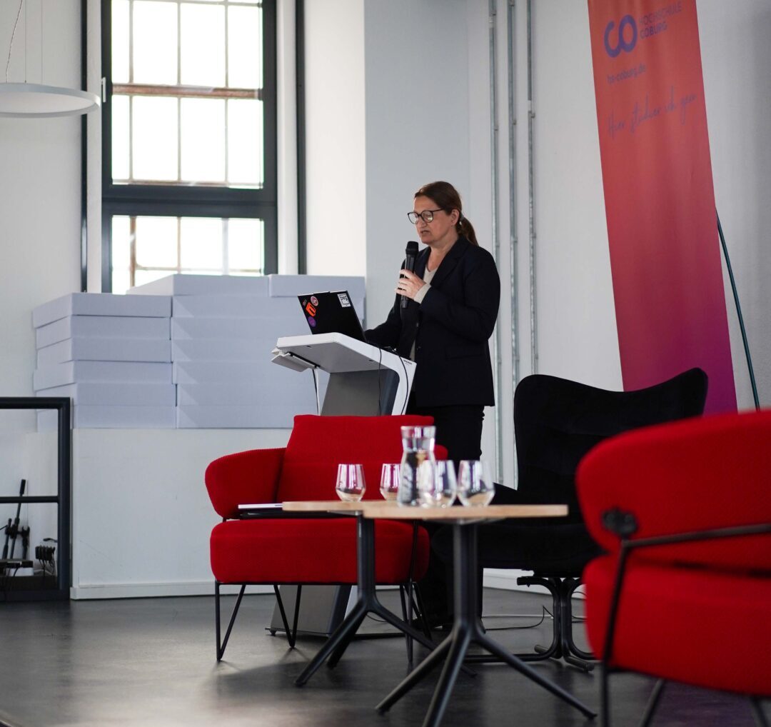 Eine Frau in Geschäftskleidung spricht in ein Mikrofon an einem Podium auf der Bühne und spricht über Themen wie Endometriose. Im Hintergrund stehen rote Stühle, ein Tisch mit Wasserflaschen und Gläsern und ein buntes Transparent. Große Fenster lassen natürliches Licht herein. at Hochschule Coburg