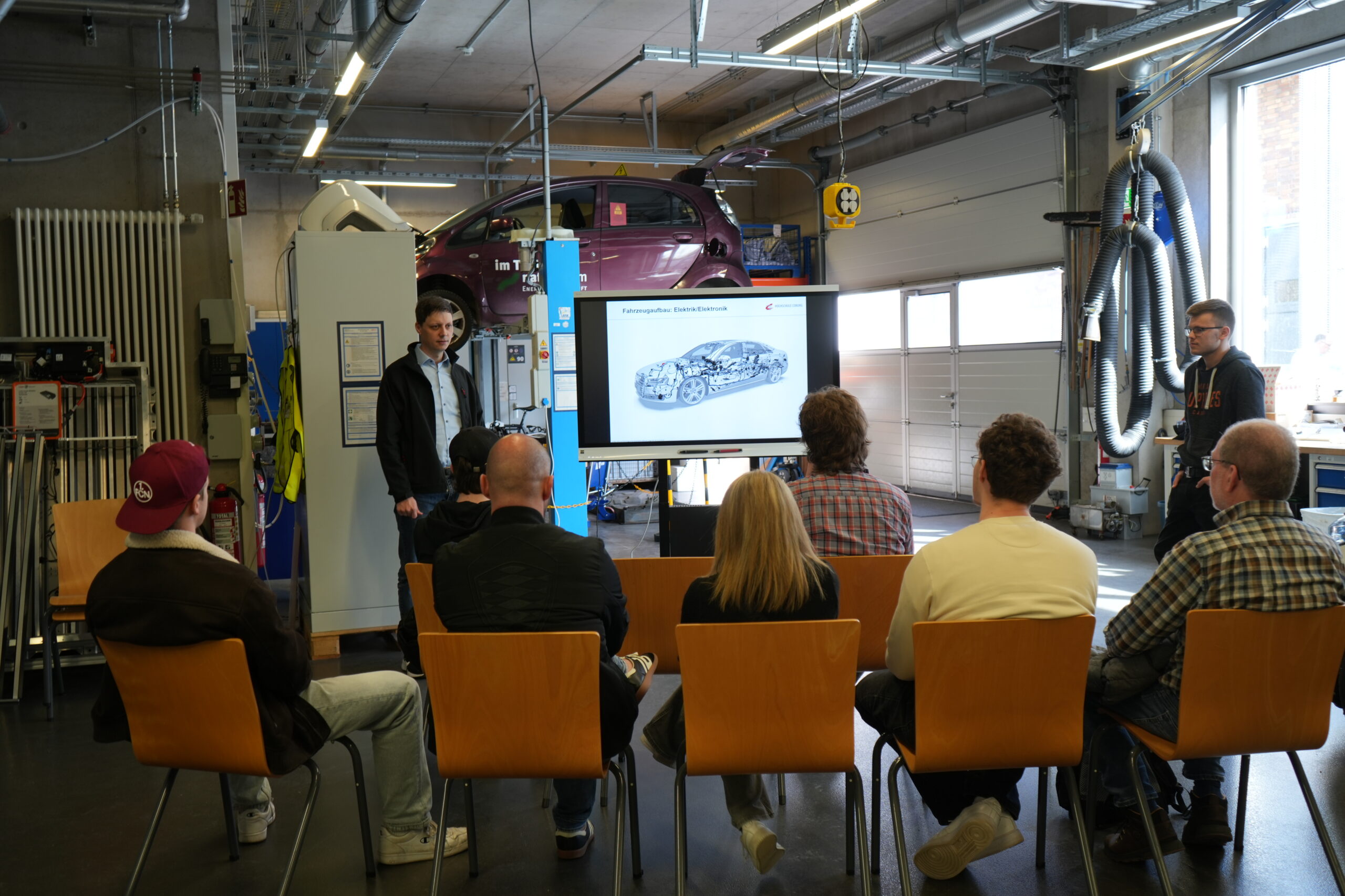 Eine Gruppe von Menschen sitzt auf Stühlen in einer Garage und sieht zu, wie ein Moderator ein Autodiagramm erklärt, das auf einem Bildschirm angezeigt wird - so wird die Campusluft eingefangen, während sie innovative Studiengänge erkunden. Im Hintergrund wird ein lila Auto auf eine Plattform gehoben. at Hochschule Coburg