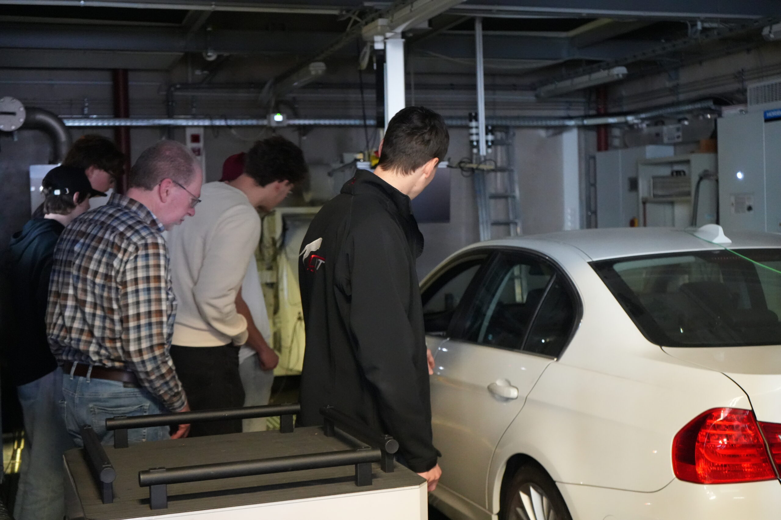 Mehrere Personen stehen um eine weiße Limousine in einer Industrie- oder Werkstattsituation auf dem Campusluft und untersuchen oder arbeiten an dem Fahrzeug bei heller Innenbeleuchtung. Im Hintergrund sind Rohre, Maschinen und Schilder zu sehen, die auf technische Studiengänge hinweisen. at Hochschule Coburg