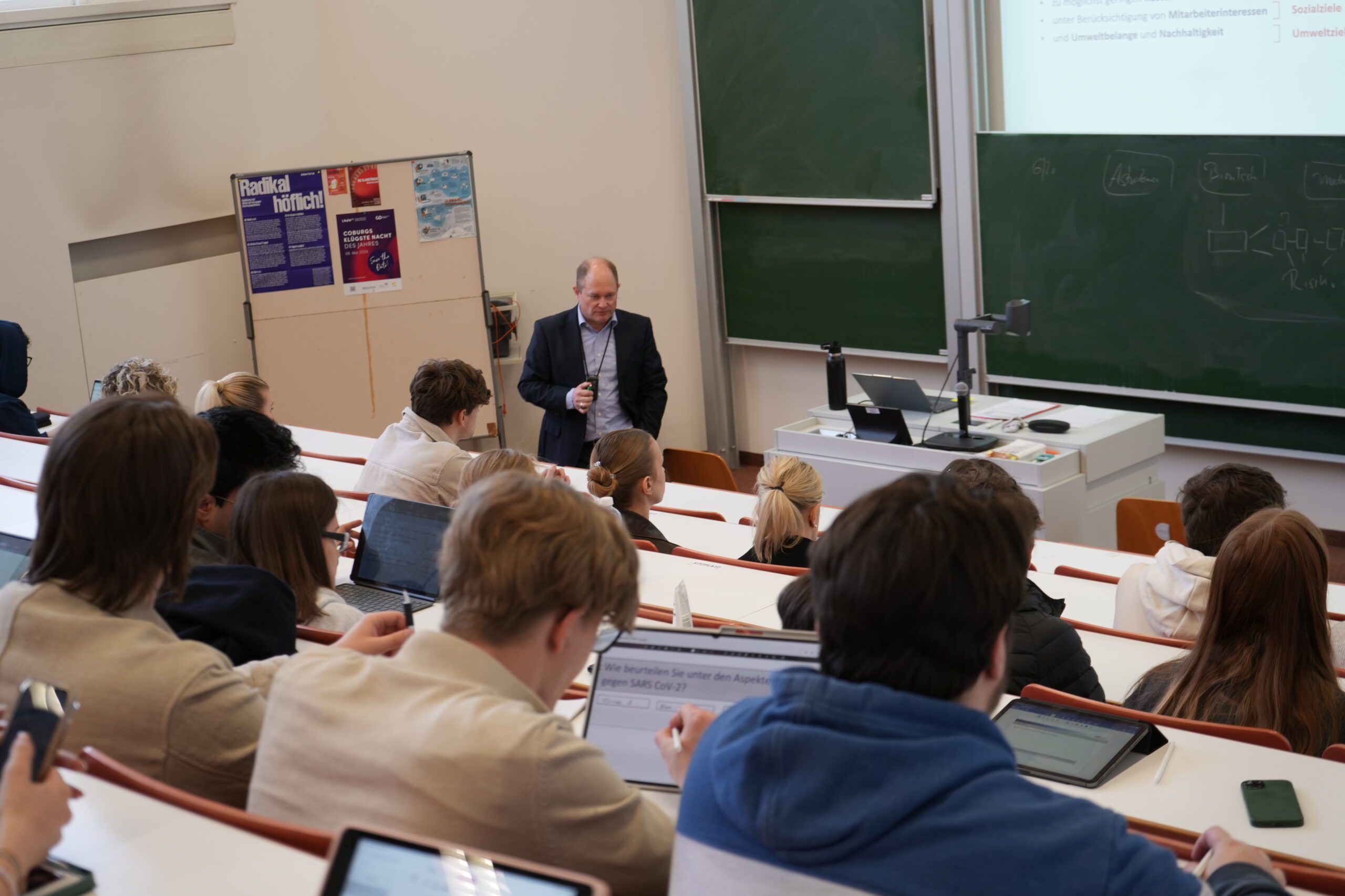 Ein Professor steht an der Stirnseite eines Universitätshörsaals und spricht zu den in Reihen sitzenden Studenten. Many erkundet die Campusluft und informiert sich über verschiedene Studiengänge, während eine Präsentation auf die Tafel projiziert wird. at Hochschule Coburg