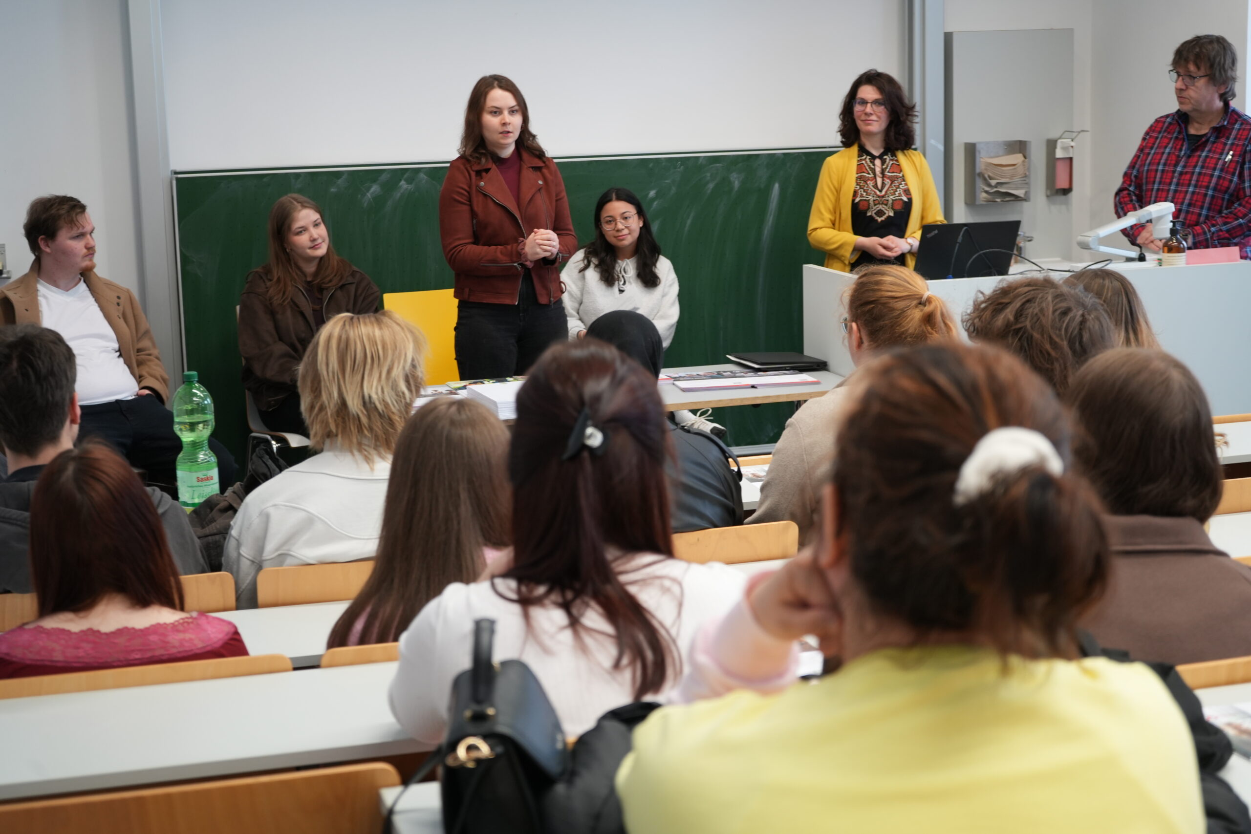 Eine Gruppe von fünf Erwachsenen steht und sitzt vorne in einem Klassenzimmer und spricht zu den in Reihen sitzenden Studenten über verschiedene Studiengänge. Die Vortragenden sind dem Publikum zugewandt, und der Raum ist von Campusluft erfüllt. Hinter ihnen sind eine Kreidetafel und ein Schreibtisch zu sehen. at Hochschule Coburg
