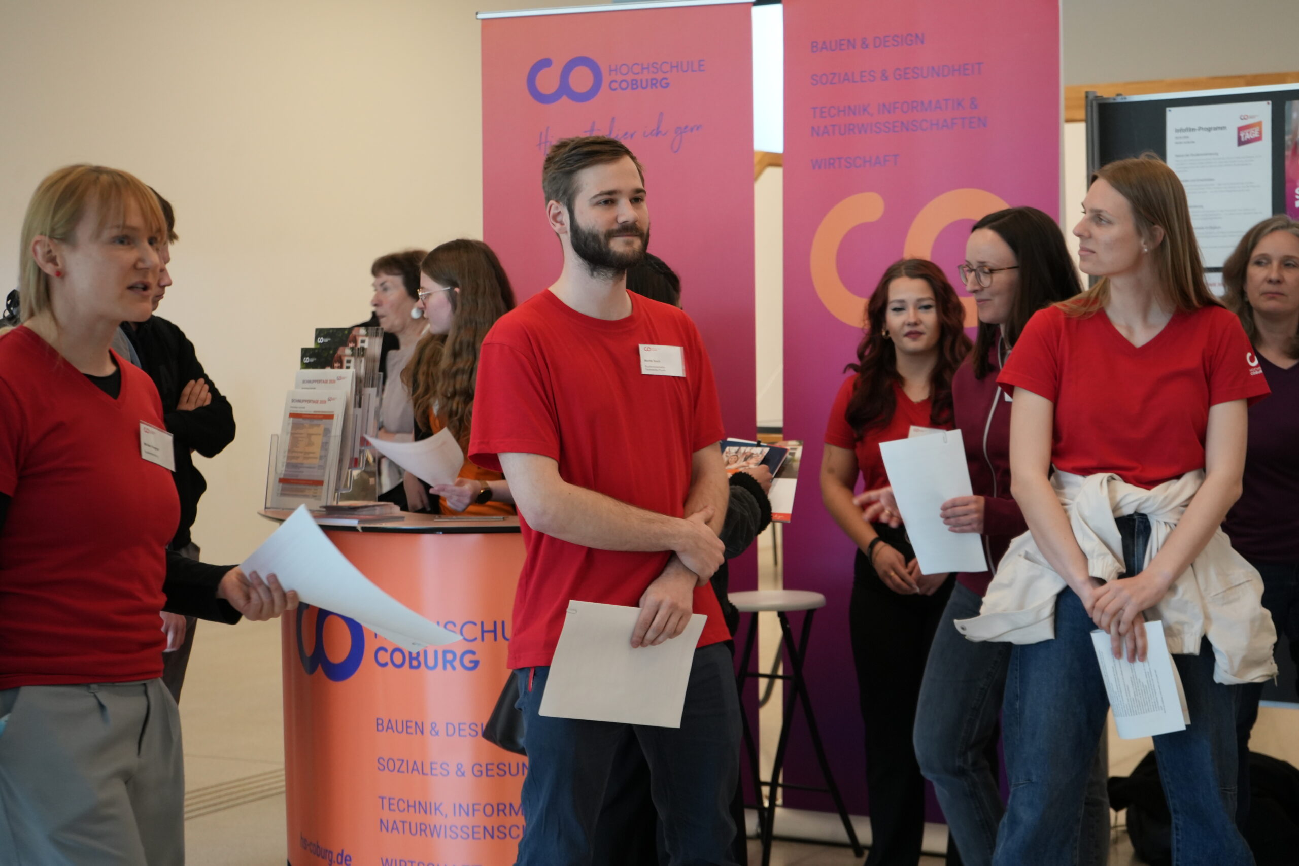 Eine Gruppe von Menschen mit roten Hemden und Namensschildern steht in einem Innenraum und hält Papiere in der Hand. Vor Bannern der Hochschule Coburg erkundet sie verschiedene Studiengänge und genießt Campusluft - und fängt die Stimmung einer Veranstaltung oder Orientierung ein. at Hochschule Coburg