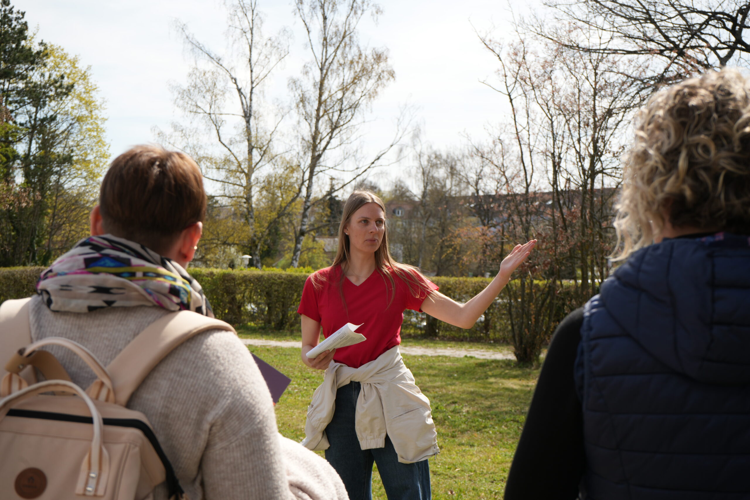 Eine Frau in einem roten Hemd gestikuliert, während sie mit zwei Personen im Freien in einer parkähnlichen Umgebung spricht, während sie gemeinsam die Campusluft erkundet und verschiedene Studiengänge diskutiert. Im sonnigen Hintergrund sind Bäume und Sträucher zu sehen. at Hochschule Coburg