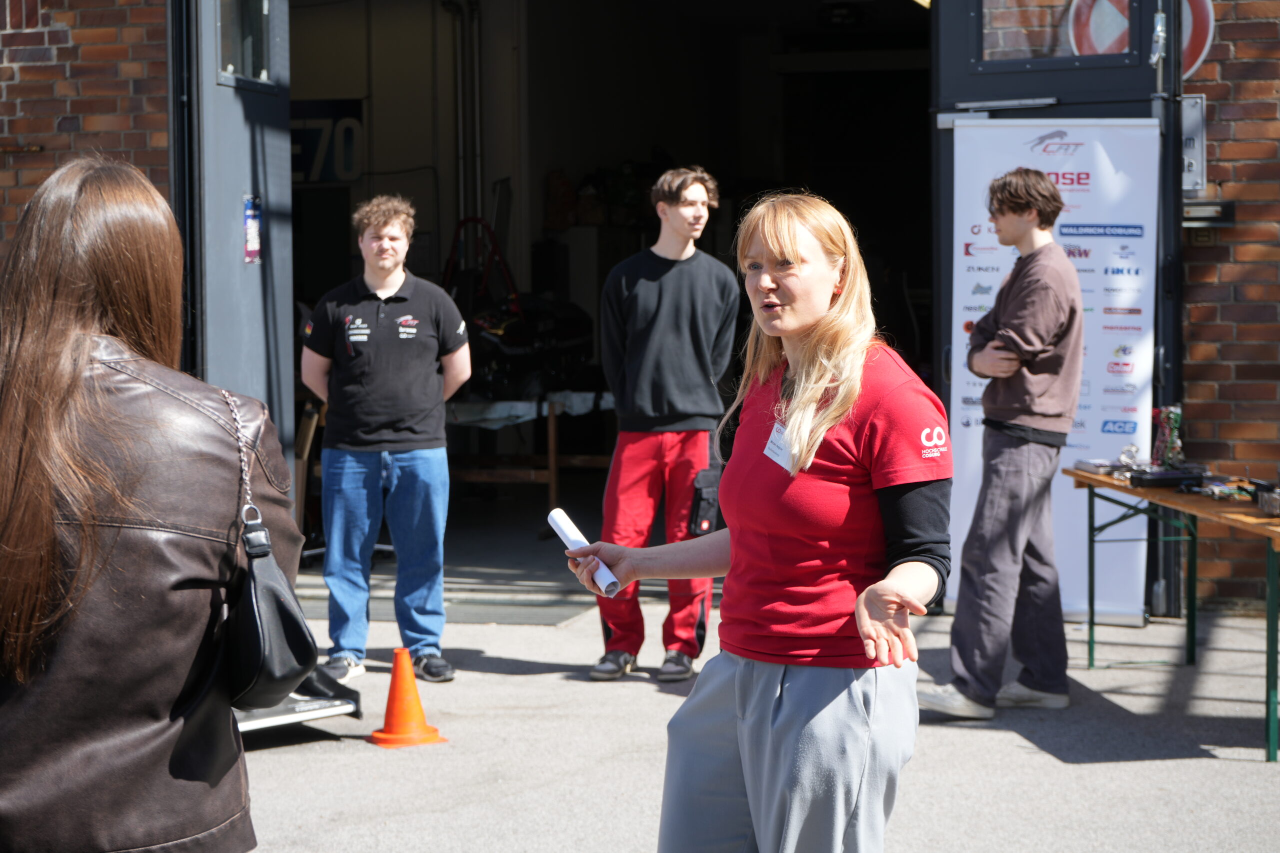 Eine Frau in einem roten Hemd spricht im Freien zu einer kleinen Gruppe, die die Campusluft genießt. Im Hintergrund diskutieren zwei Personen in der Nähe einer Garageneinfahrt und eines aufrollbaren Banners über Studiengänge, während das Sonnenlicht Schatten auf den Boden wirft. at Hochschule Coburg