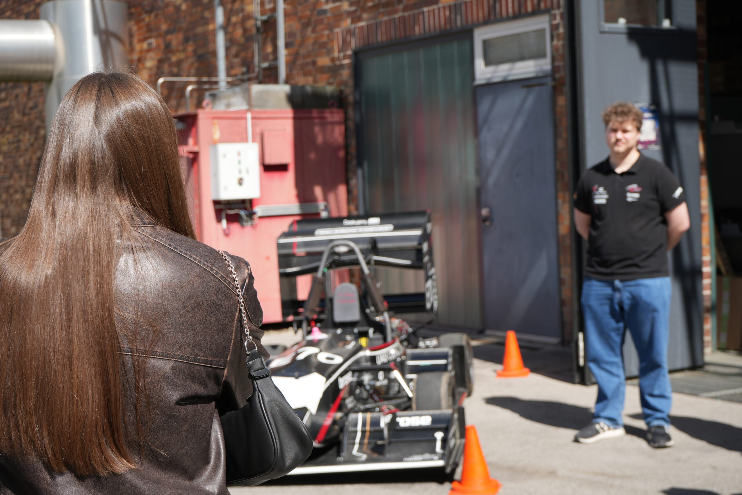 Eine Frau mit langen braunen Haaren steht einem Mann gegenüber, der neben einem schwarz-weißen Rennwagen steht, der vor einem Backsteingebäude geparkt ist und die Campusluft einfängt. Orangefarbene Verkehrskegel umgeben das Auto, und der Mann trägt ein schwarzes Hemd und blaue Jeans. at Hochschule Coburg