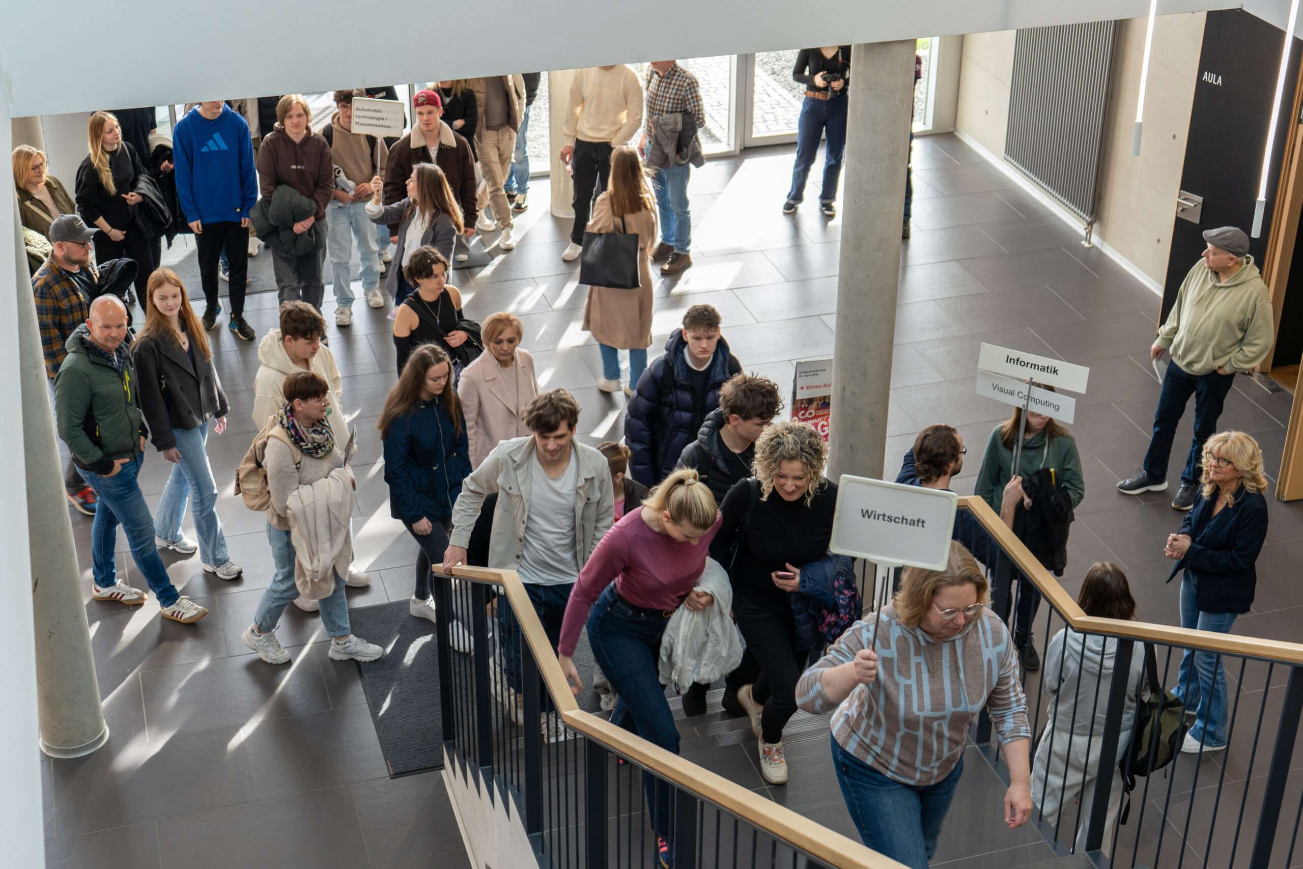 Eine große Gruppe von Menschen, zumeist junge Erwachsene, versammelt sich im Innenbereich, um Studiengänge zu erkunden. Einige folgen den Führern, die Schilder mit der Aufschrift "Wirtschaft" und "Informatik" halten, während andere die Campusluft genießen, während sie die Treppe hinaufgehen oder sich im Hauptgeschoss unterhalten. at Hochschule Coburg