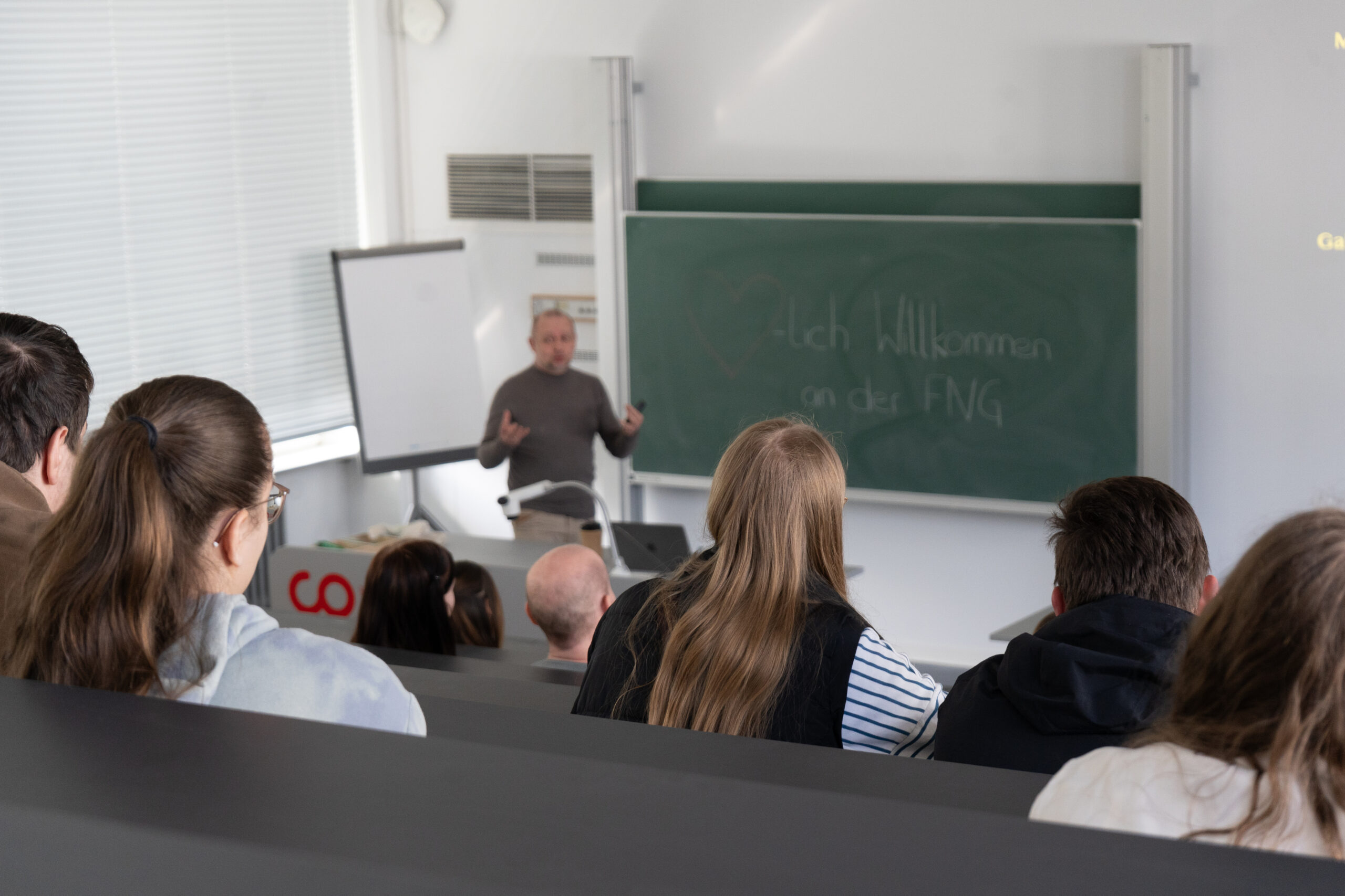 Ein Dozent spricht zu Studenten in einem Klassenzimmer mit Reihenbestuhlung. Auf der Kreidetafel hinter ihm steht "Herzlich Willkommen an der FNG". Mehrere Studenten, die die Campusluft und die neuen Studiengänge kennenlernen wollen, hören aufmerksam zu und schauen dem Dozenten zu. at Hochschule Coburg