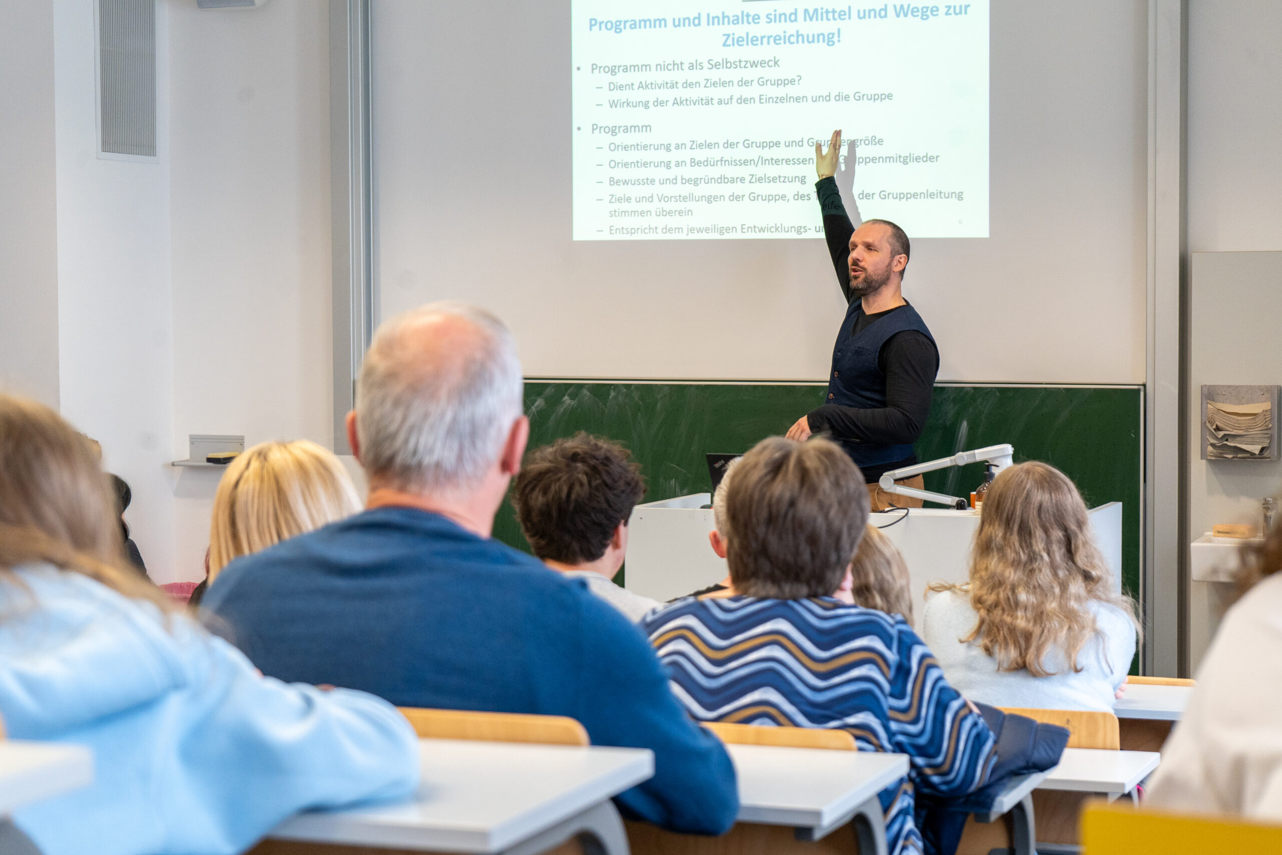 Ein Dozent steht vorne in einem Klassenzimmer und zeigt auf eine Präsentationsfolie in deutscher Sprache über verschiedene Studiengänge, während die Studierenden inmitten von Tischen, Stühlen und einer Kreidetafel sitzen und aufmerksam zuhören und die echte Campusluft genießen. at Hochschule Coburg