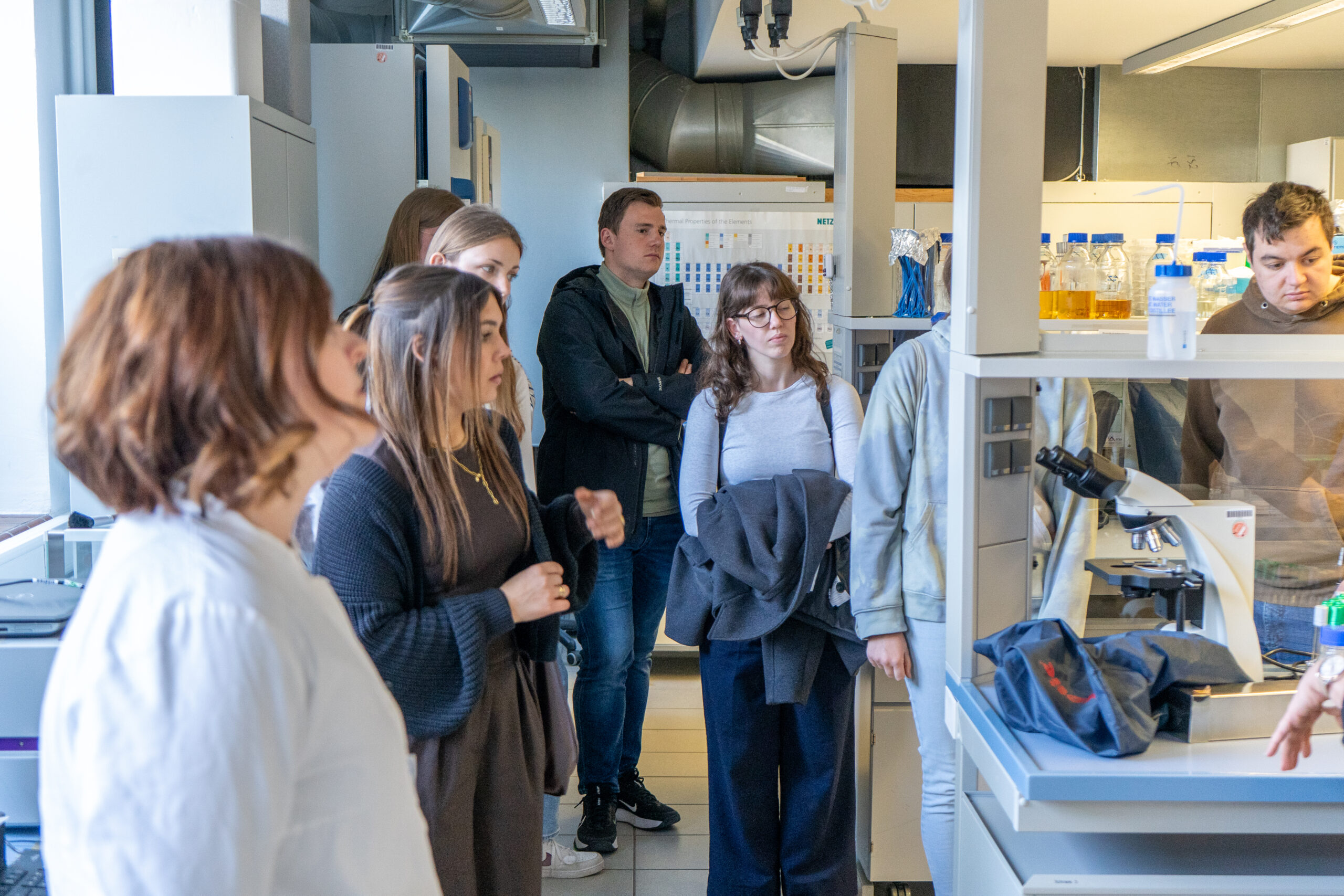 Eine Gruppe von Menschen steht in einem Labor und hört aufmerksam zu, wenn jemand aus dem Off die Campusluft erkundet und mehr über verschiedene Studiengänge erfährt. Laborgeräte, Regale und Flaschen umgeben sie, einige halten Jacken oder Taschen. at Hochschule Coburg