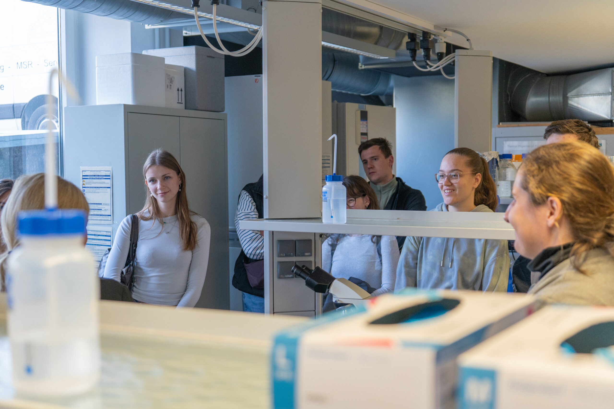 Eine Gruppe junger Leute steht in einem hellen Labor zusammen, einige lächeln und hören aufmerksam zu. Die Campusluft ist lebendig, Laborgeräte und Flaschen füllen den Vordergrund und spiegeln ihre Begeisterung für innovative Studiengänge wider. at Hochschule Coburg