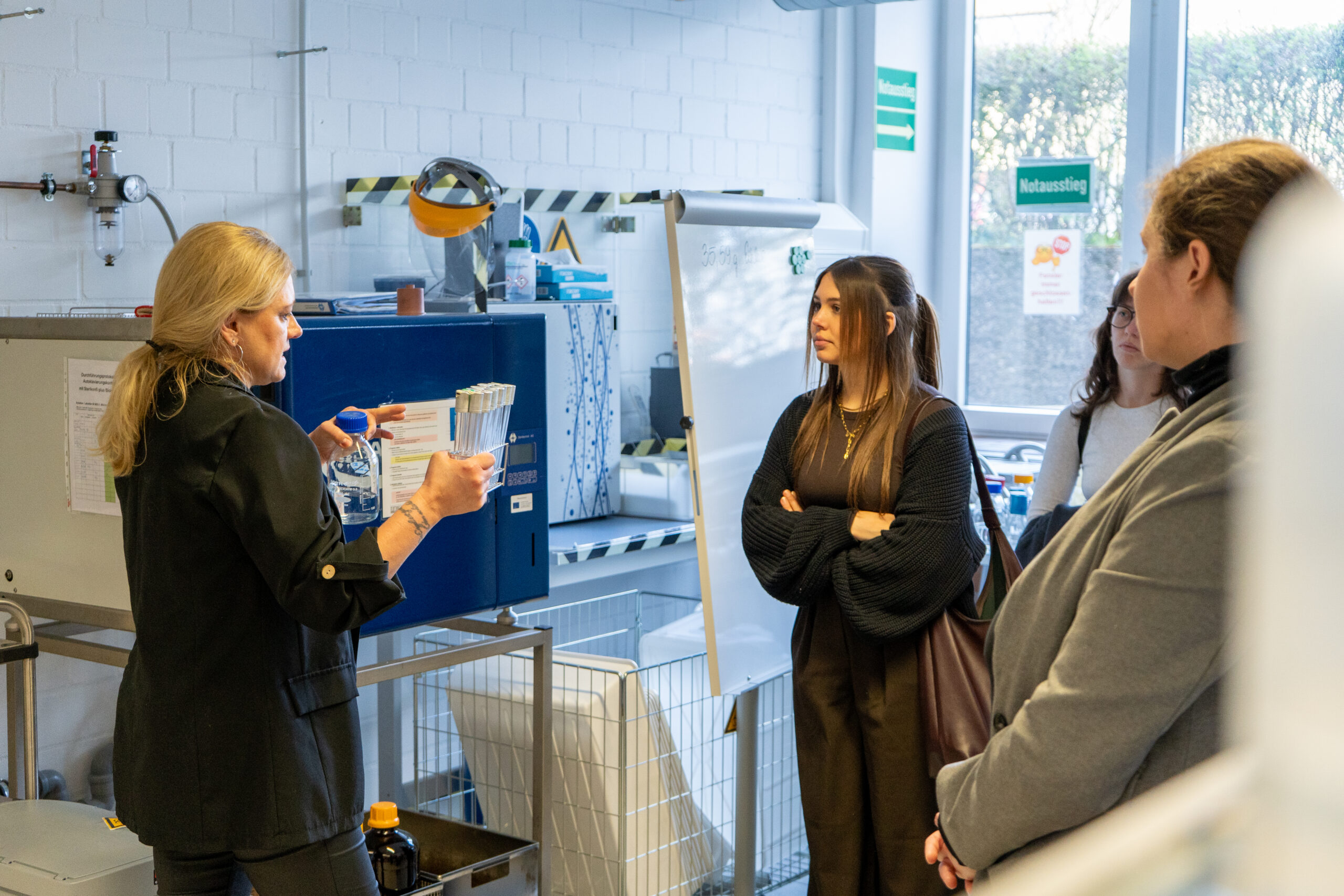 Eine Frau erklärt oder demonstriert drei Personen in einer Laborumgebung etwas, wobei sie Röhrchen oder Behälter hält. Im Hintergrund sind Geräte, Sicherheitsschilder und die lebendige Campusluft verschiedener Studiengänge zu sehen. at Hochschule Coburg