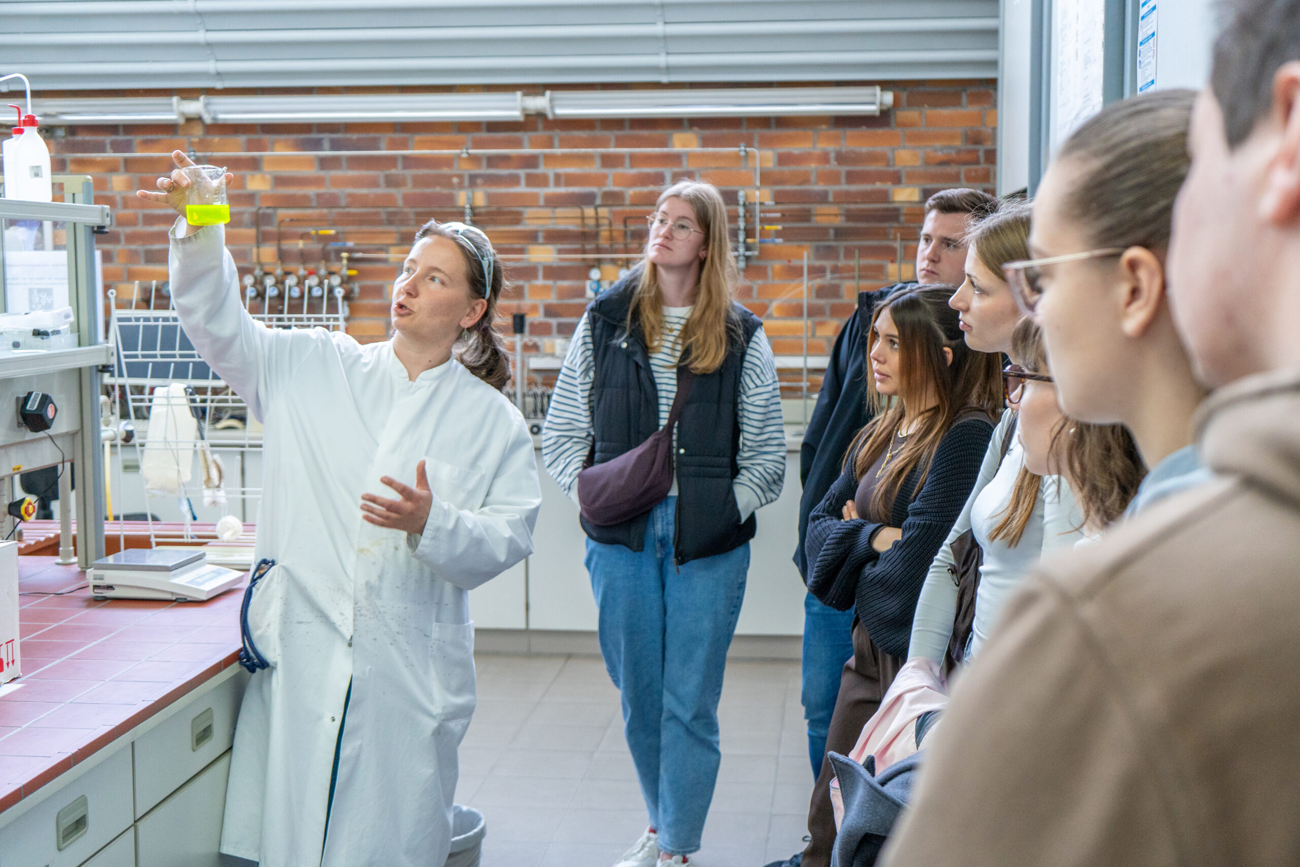 Ein Wissenschaftler in einem weißen Laborkittel hält ein Becherglas mit einer gelben Flüssigkeit in die Höhe und erklärt den aufmerksamen Studenten, die eifrig dabei sind, verschiedene Studiengänge zu erkunden und echte Campusluft im Labor zu erleben, deren Eigenschaften. at Hochschule Coburg