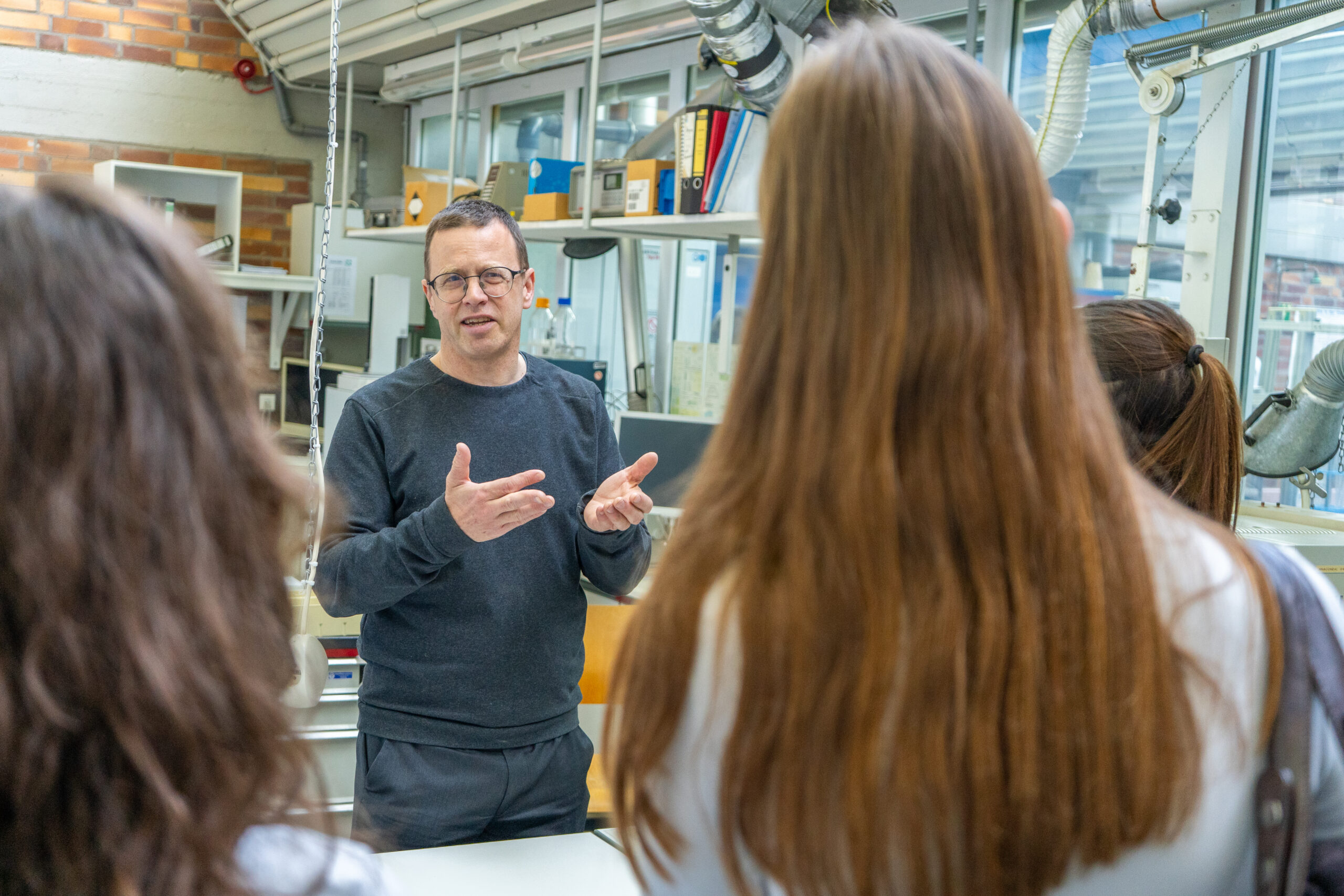 Ein Mann mit Brille gestikuliert, während er mit drei Personen mit langen Haaren in einem hell erleuchteten Labor oder Klassenzimmer spricht, wo sie gemeinsam Campusluft schnuppern und spannende Studiengänge erkunden. Regale, Bücher und Geräte füllen den Raum. at Hochschule Coburg