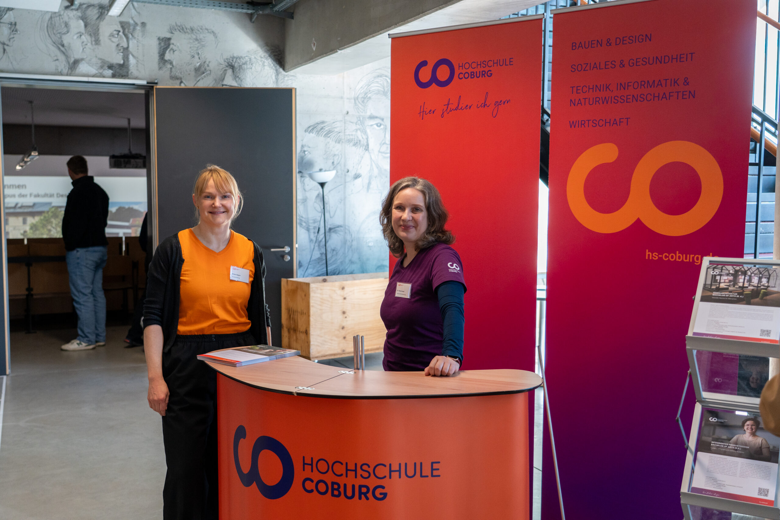 Zwei Frauen stehen bei einer Indoor-Veranstaltung hinter einem orange-roten Infostand der Hochschule Coburg. Auf Bannern ist der Name der Hochschule zu lesen, rechts daneben liegen Broschüren über Campusluft und Studiengänge aus. at Hochschule Coburg