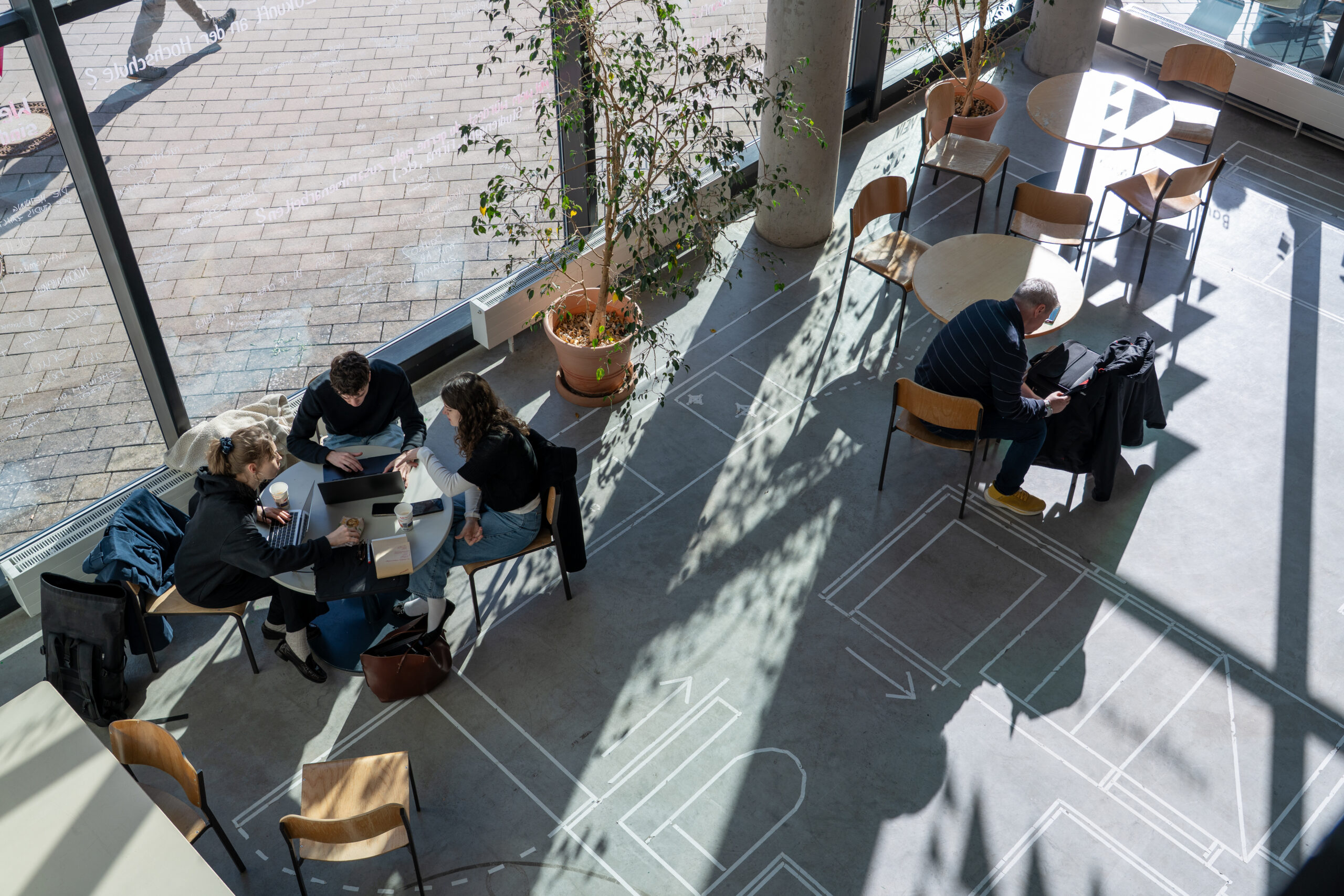 Draufsicht auf mehrere Personen, die an Tischen in einem sonnendurchfluteten Café oder Lounge-Bereich sitzen und die Campusluft durch große Fenster und Pflanzen genießen. Einige erkundet neue Studiengänge zusammen, während eine Person allein mit einem Laptop inmitten geometrischer Bodenmuster sitzt. at Hochschule Coburg