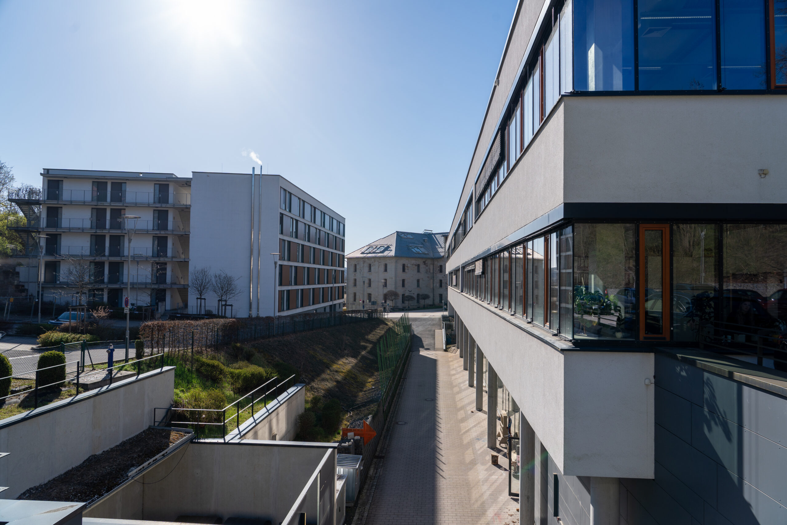 Eine sonnige Tagesansicht von modernen Bürogebäuden mit großen Fenstern, einem Gehweg dazwischen und einem älteren Steingebäude im Hintergrund fängt die lebendige Campusluft unter einem klaren blauen Himmel ein. at Hochschule Coburg