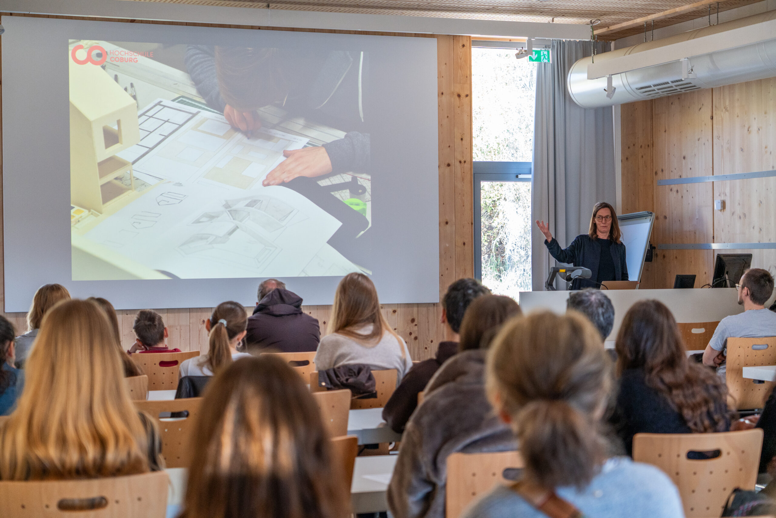 Eine Frau hält in einem Klassenzimmer mit sitzenden Studenten einen Vortrag über architektonische Studiengänge. Auf der Leinwand sind Modelle zu sehen, während die Studenten in einem holzgetäfelten Raum Campusluft schnuppern, ganz auf die Vortragende konzentriert. at Hochschule Coburg