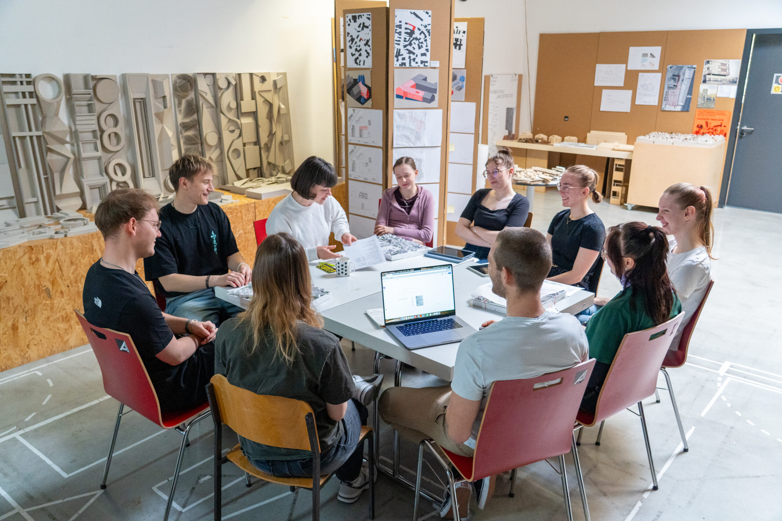 Neun Personen sitzen um einen Tisch in einem Klassenzimmer oder Atelier und diskutieren oder arbeiten zusammen. Laptops, Papiere und Modelle liegen auf dem Tisch; Architekturmodelle und Zeichnungen säumen die Wände - die Campusluft ist spürbar, während gemeinsame Studiengänge erkundet werden. at Hochschule Coburg