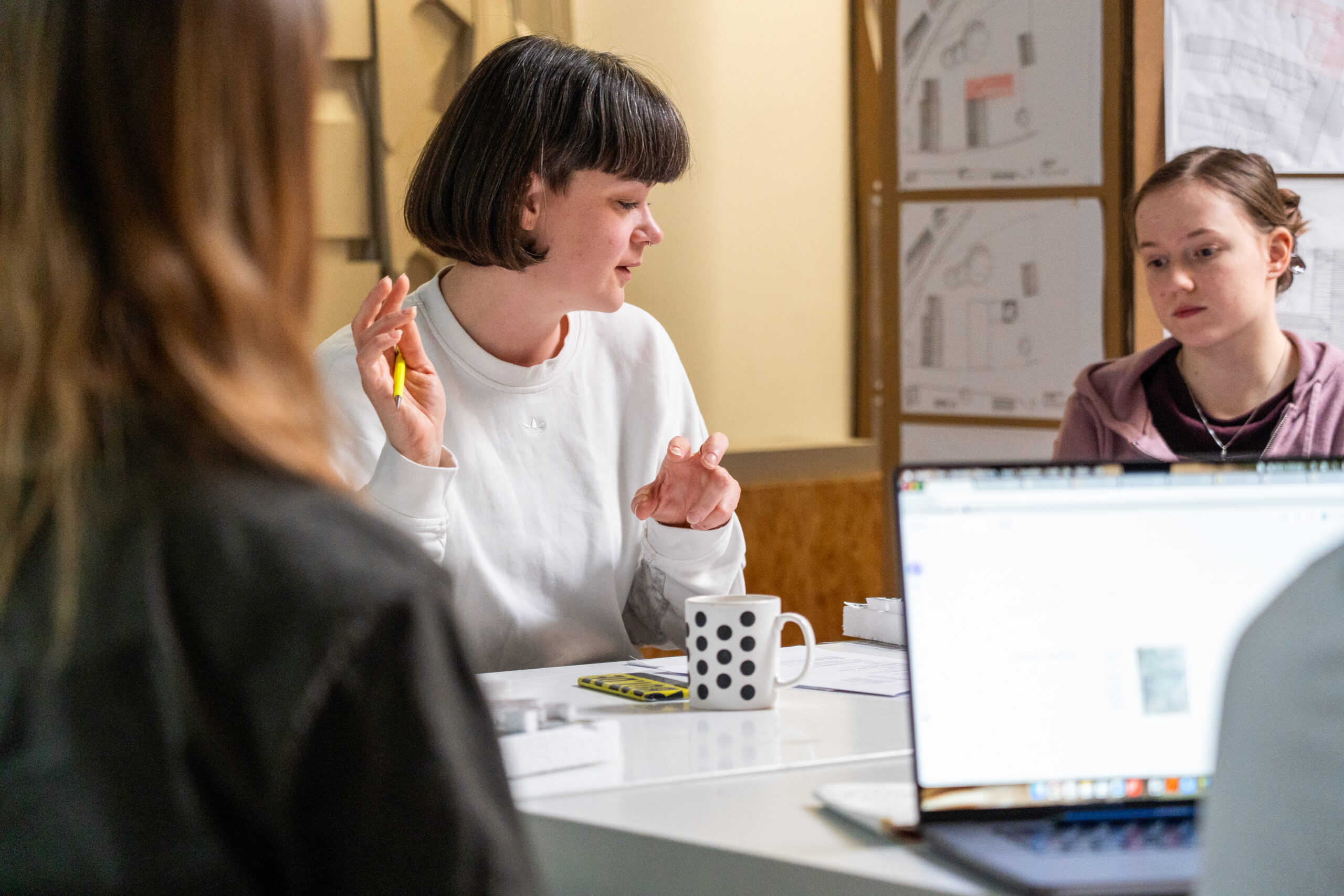 Eine Frau mit kurzen Haaren gestikuliert während einer Besprechung an einem Tisch mit Papieren, einem Becher und einem Laptop; zwei andere hören aufmerksam zu. Architektonische Zeichnungen an der Wand weisen auf Möglichkeiten hin, verschiedene Studiengänge zu erkunden und Campusluft zu schnuppern. at Hochschule Coburg