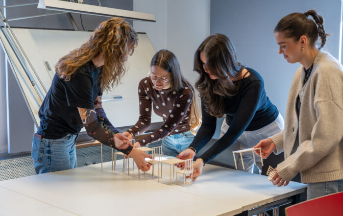 Vier Frauen stehen um einen Tisch und arrangieren gemeinsam kleine Architekturmodelle von Gebäuden. Im Hintergrund steht eine Staffelei, die die lebendige Campusluft der kreativen Studiengänge in diesem Bildungsarbeitsraum einfängt. at Hochschule Coburg