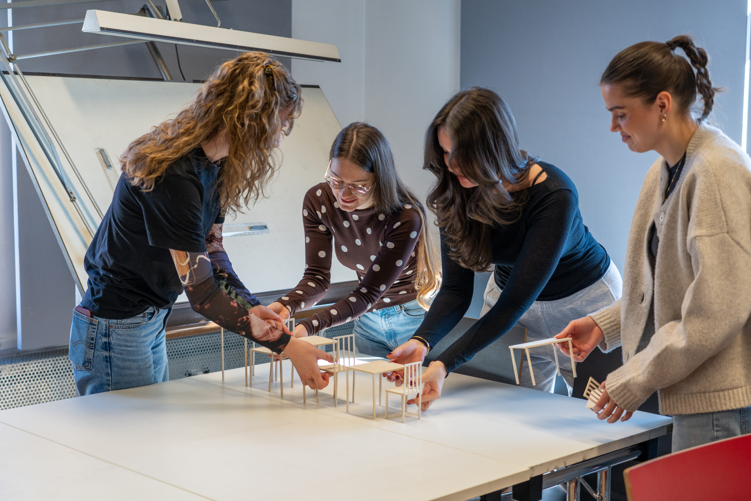 Vier Frauen stehen um einen Tisch und arrangieren gemeinsam kleine Architekturmodelle von Gebäuden. Im Hintergrund steht eine Staffelei, die die lebendige Campusluft der kreativen Studiengänge in diesem Bildungsarbeitsraum einfängt. at Hochschule Coburg