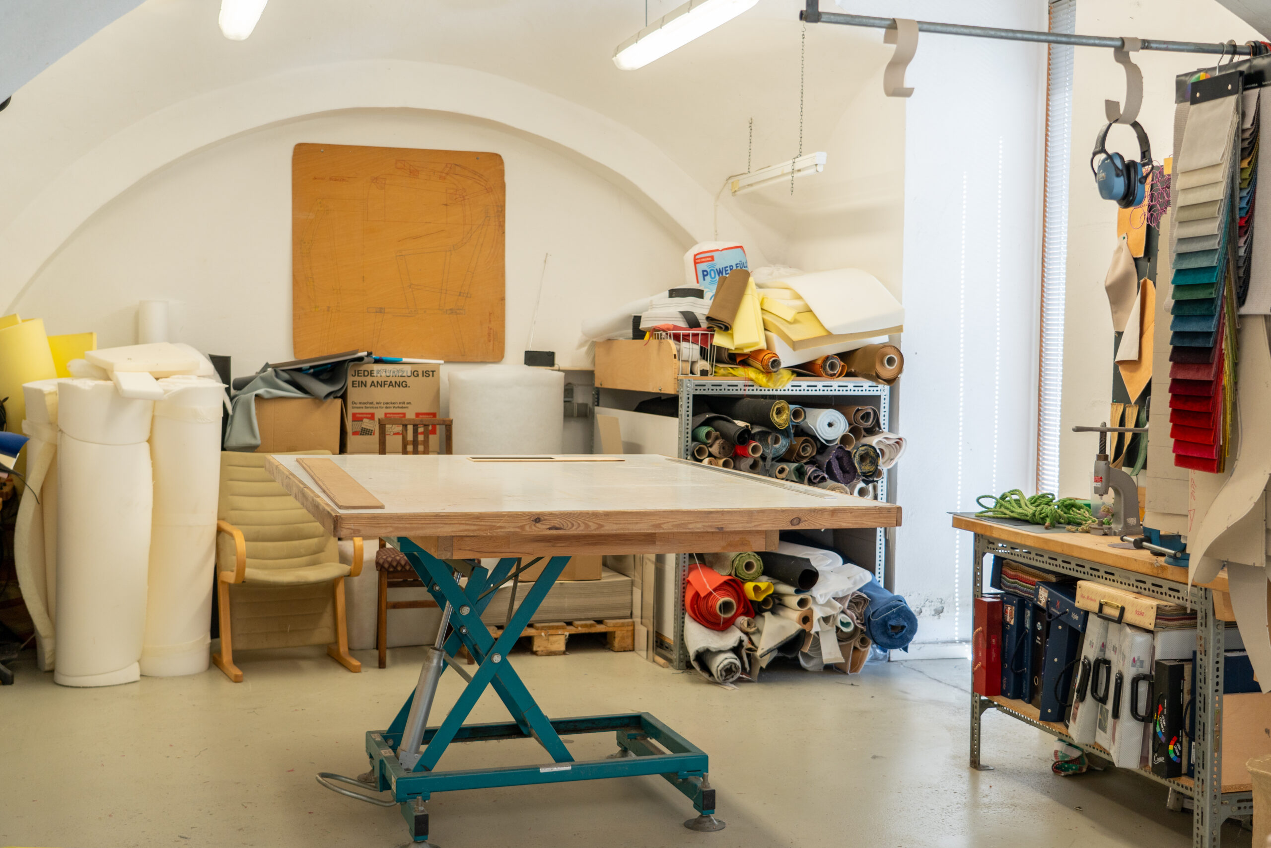 Eine Werkstatt mit einem zentralen hölzernen Arbeitstisch, Regalen mit Stoff- und Schaumstoffrollen, an den Wänden hängenden Werkzeugen und einem Stuhl in der Ecke unter hellen Oberlichtern - perfekt für kreative Studiengänge in einer inspirierenden Campusluft. at Hochschule Coburg