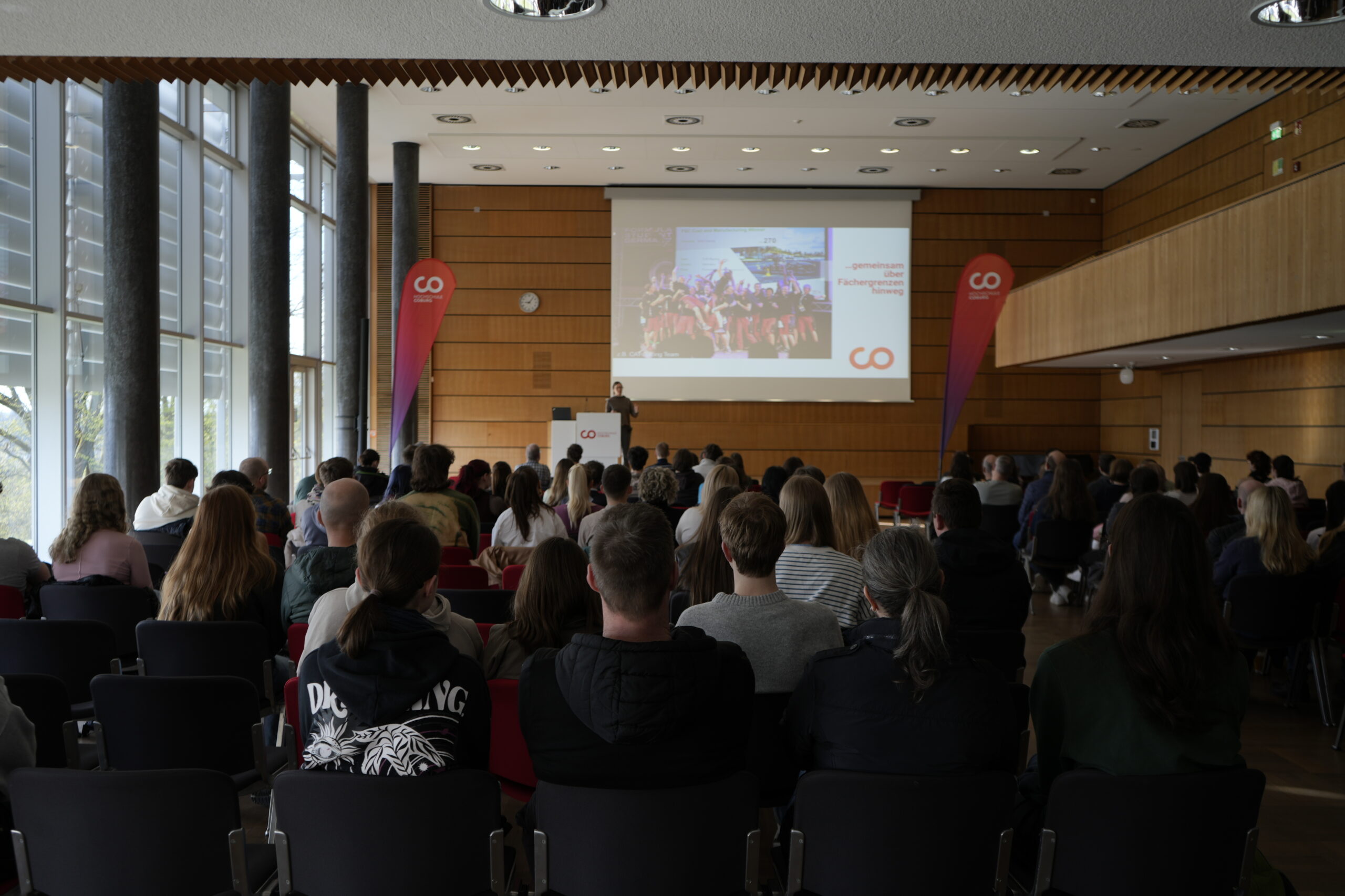 In einem modernen, gut beleuchteten Konferenzraum mit hohen Fenstern und Holzwänden sitzt eine Gruppe von Personen einem Redner gegenüber, der Folien auf einem großen Bildschirm präsentiert, während sie die Campusluft und spannende Studiengänge erkundet. Zwei vertikale Banner mit "GO"-Logos stehen neben dem Bildschirm. at Hochschule Coburg