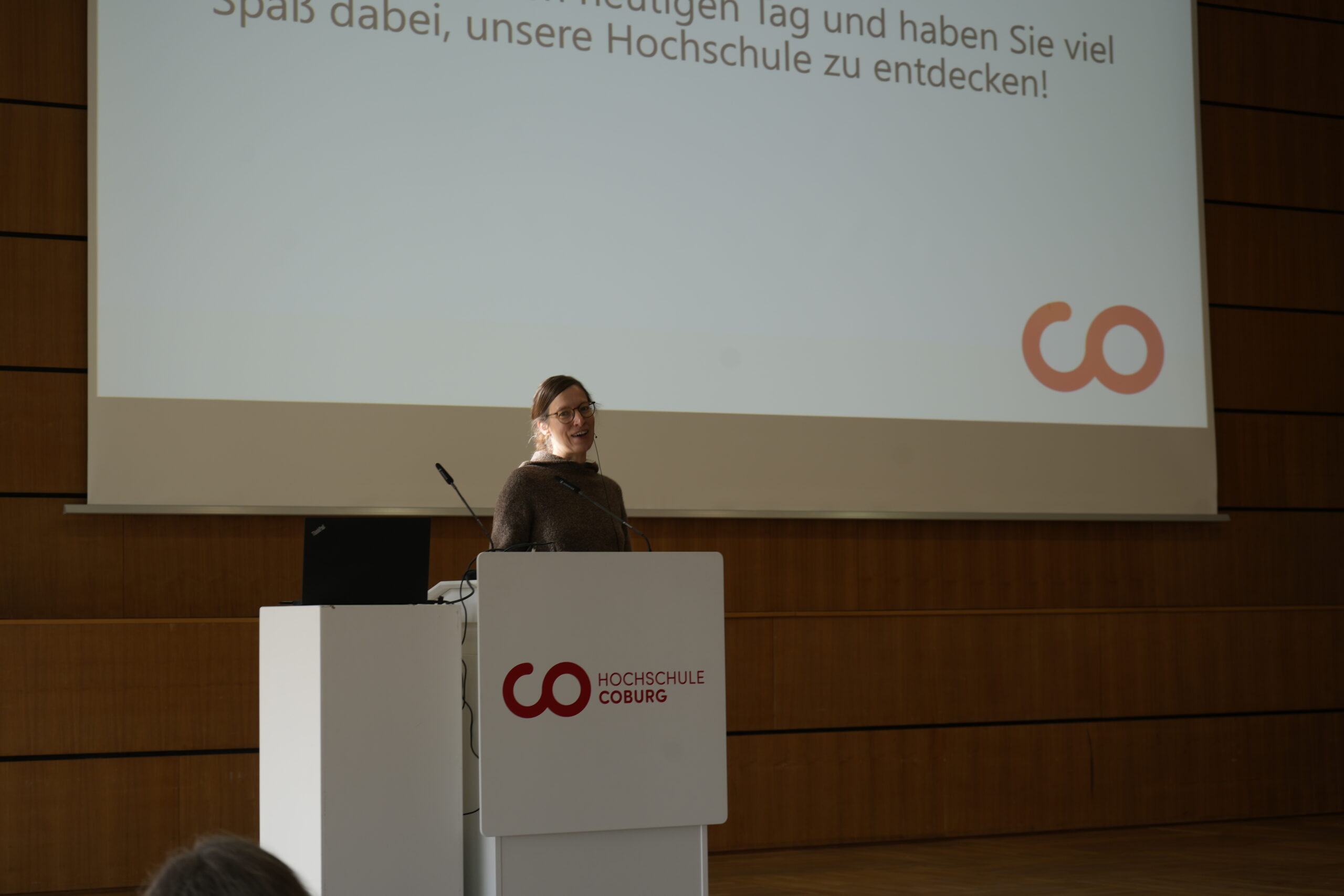 Eine Frau steht an einem Podium und hält einen Vortrag in einem Hörsaal der Hochschule Coburg. Die Folie hebt den deutschen Text und ein rotes, kreisförmiges Logo hervor und stellt verschiedene Studiengänge vor, die angeboten werden, und fängt die lebendige Campusluft ein. at Hochschule Coburg