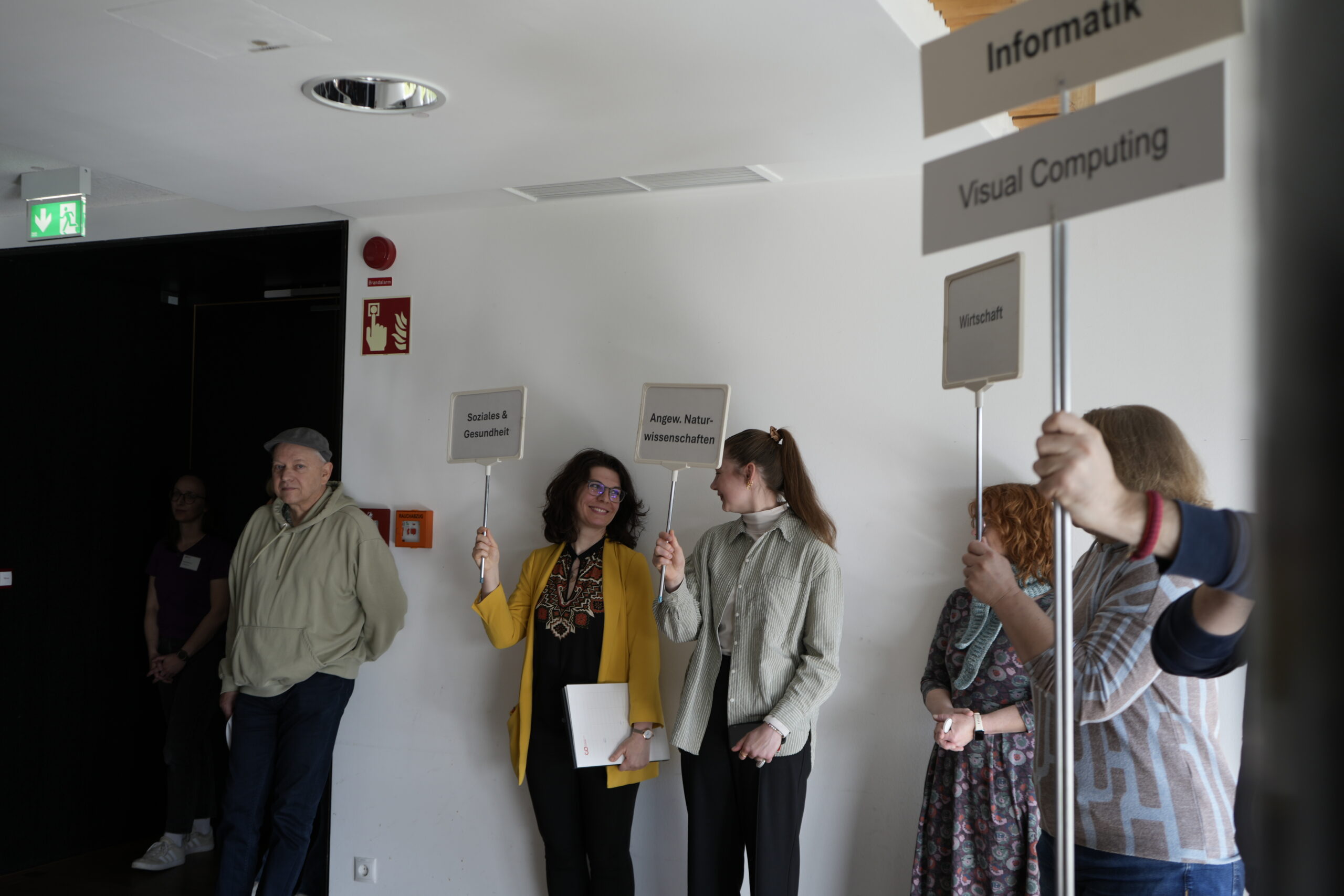 Eine Gruppe von Menschen steht im Innenraum, einige halten Schilder mit Aufschriften wie "Soziales & Gesundheit", "Informatik" und "Visual Computing". Zwei Frauen in der Nähe der Mitte lächeln und unterhalten sich, während sie gemeinsam Studiengänge erkunden und Campusluft schnuppern. at Hochschule Coburg