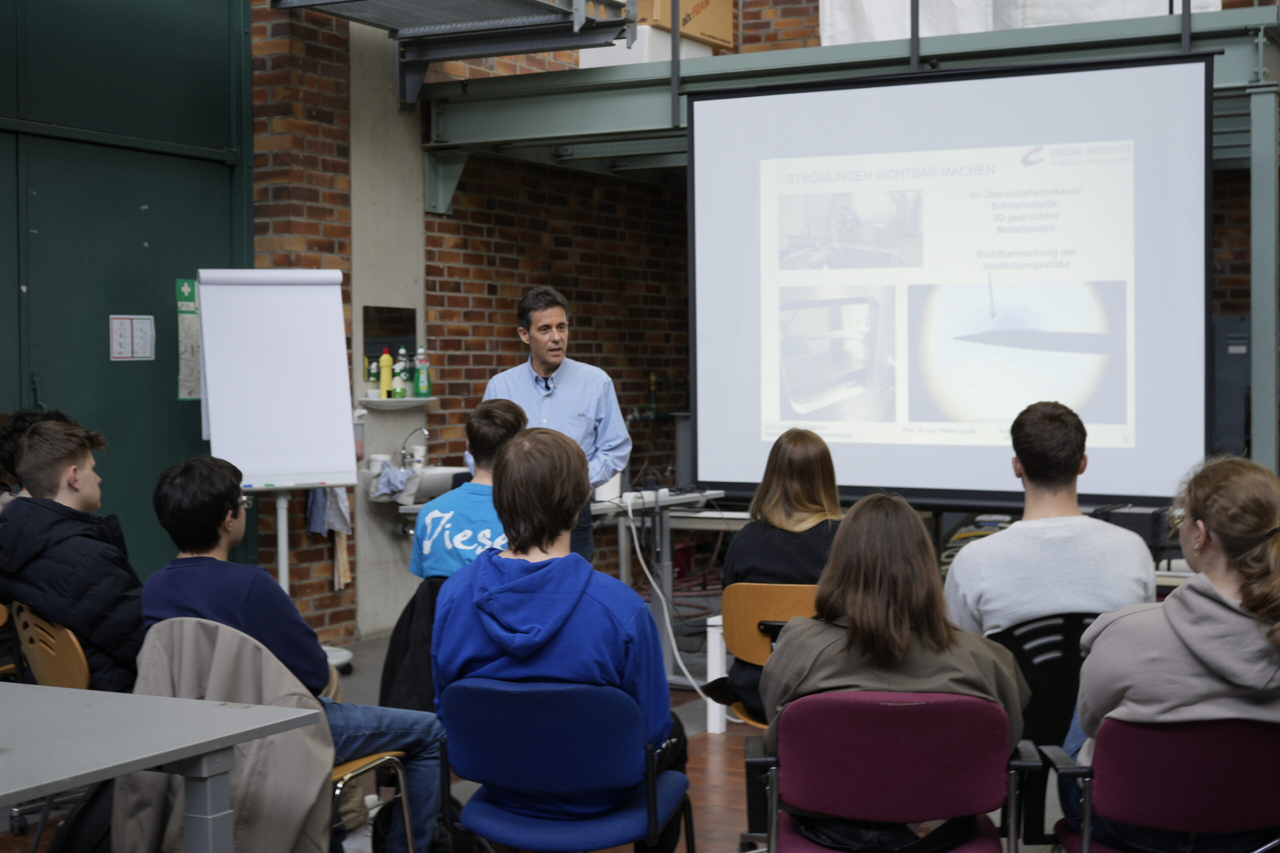 Ein Mann steht und präsentiert vor einer sitzenden Gruppe in einem Klassenzimmer, neben ihm ein Bildschirm mit Dias und ein Flipchart - perfekt für alle, die Campusluft schnuppern oder verschiedene Studiengänge erkundet haben. Der Raum hat Backsteinwände und industrielle Elemente. at Hochschule Coburg