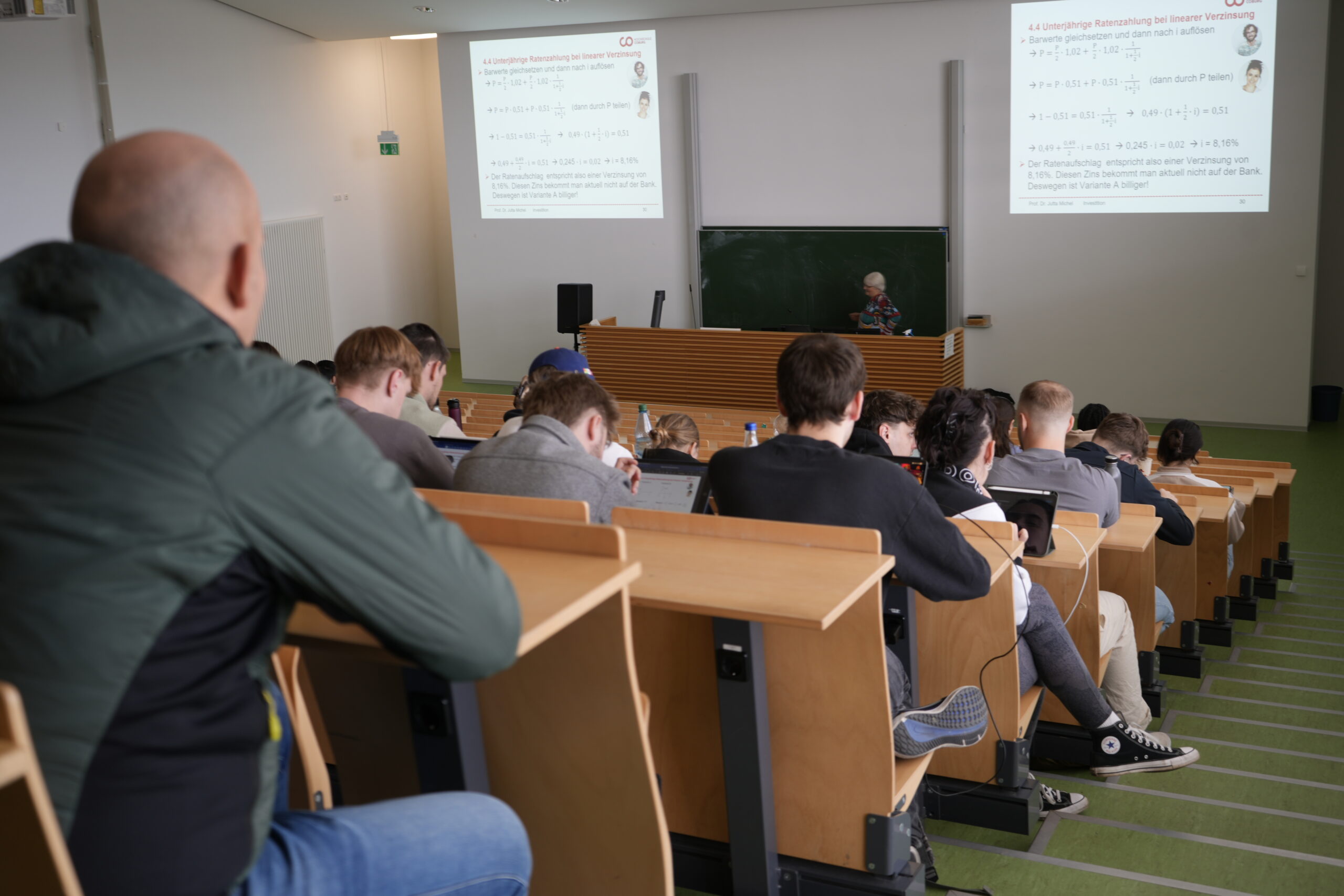 Die Studenten sitzen an gestuften Holzpulten und genießen die Campusluft, während sie einem Dozenten gegenübersitzen, der mathematische Formeln auf projizierten Folien präsentiert. Die Szene fängt die Essenz verschiedener Studiengänge in einem akademischen Klassenzimmer ein. at Hochschule Coburg