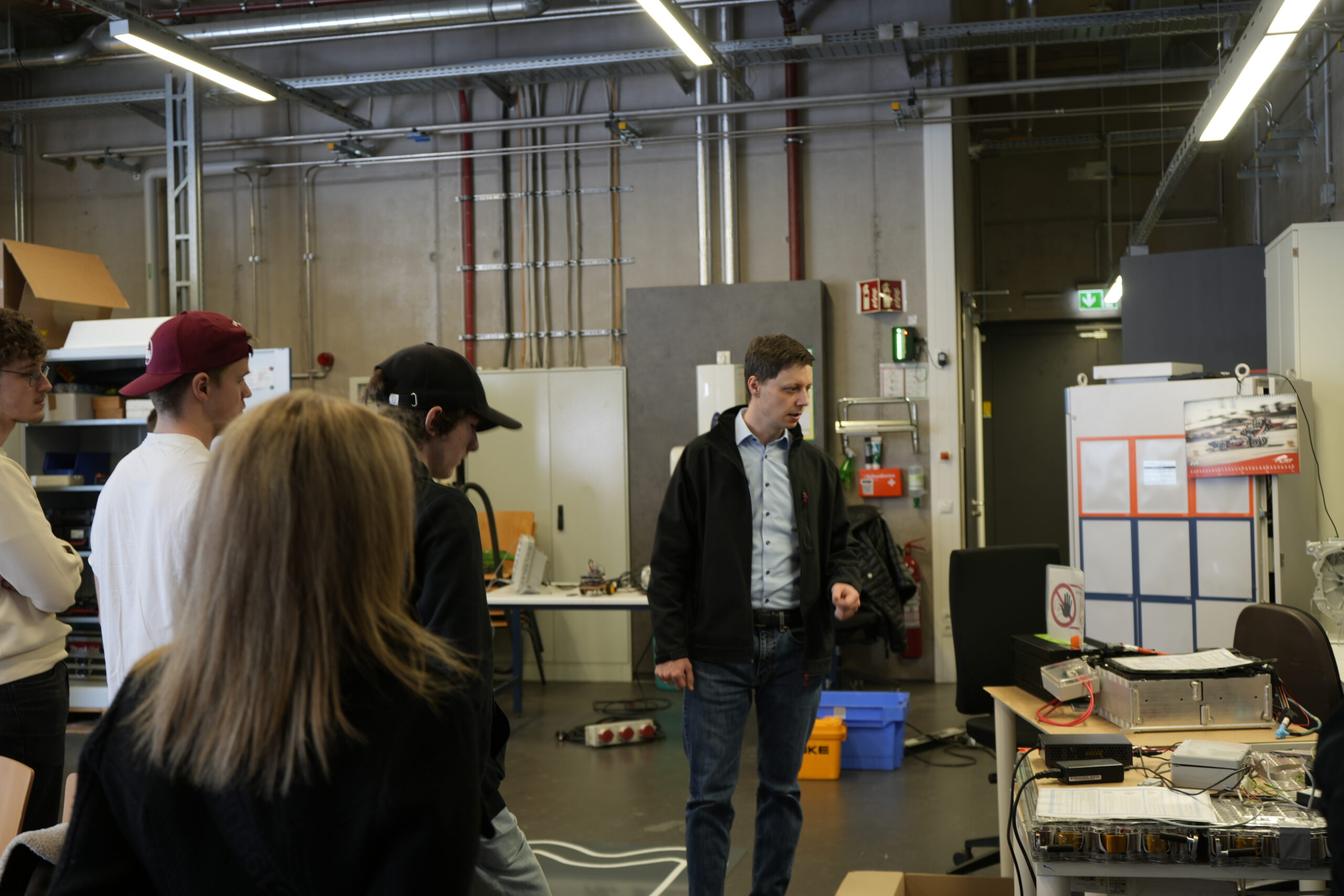 Ein Mann in einer Jacke spricht zu vier Personen in einer Werkstatt oder einem Labor, umgeben von Geräten und Kabeln. Die Gruppe, die in Campusluft getaucht ist, hört zu, während er technische Details erklärt - vielleicht diskutiert er über innovative Studiengänge. at Hochschule Coburg