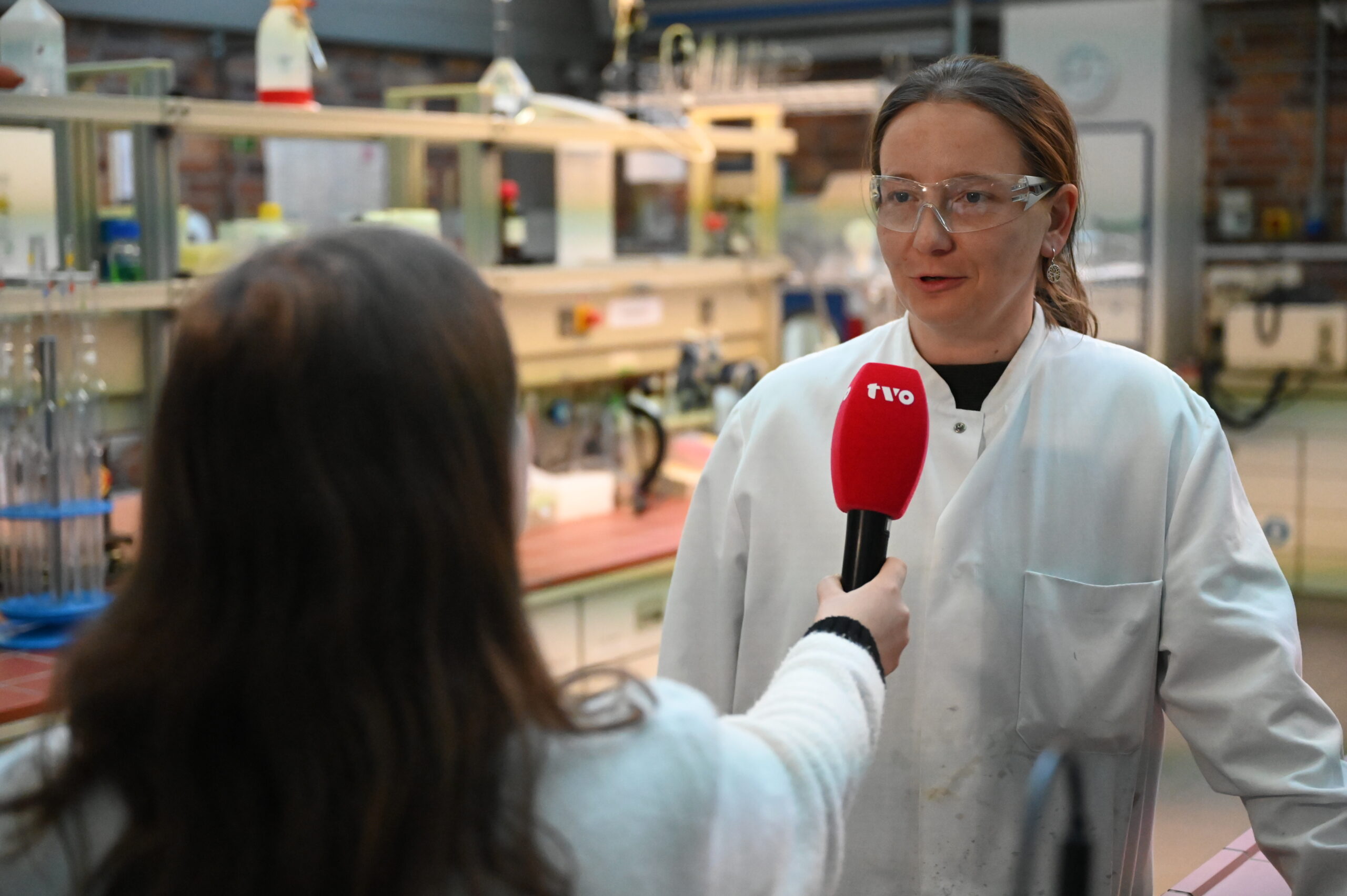Eine Frau im Laborkittel und mit Schutzbrille wird von einer anderen Frau interviewt, die ein rotes "rvo"-Mikrofon hält und den TVO-Zuschauern einen Einblick in das Laborleben zwischen Regalen und wissenschaftlichen Geräten bietet. at Hochschule Coburg