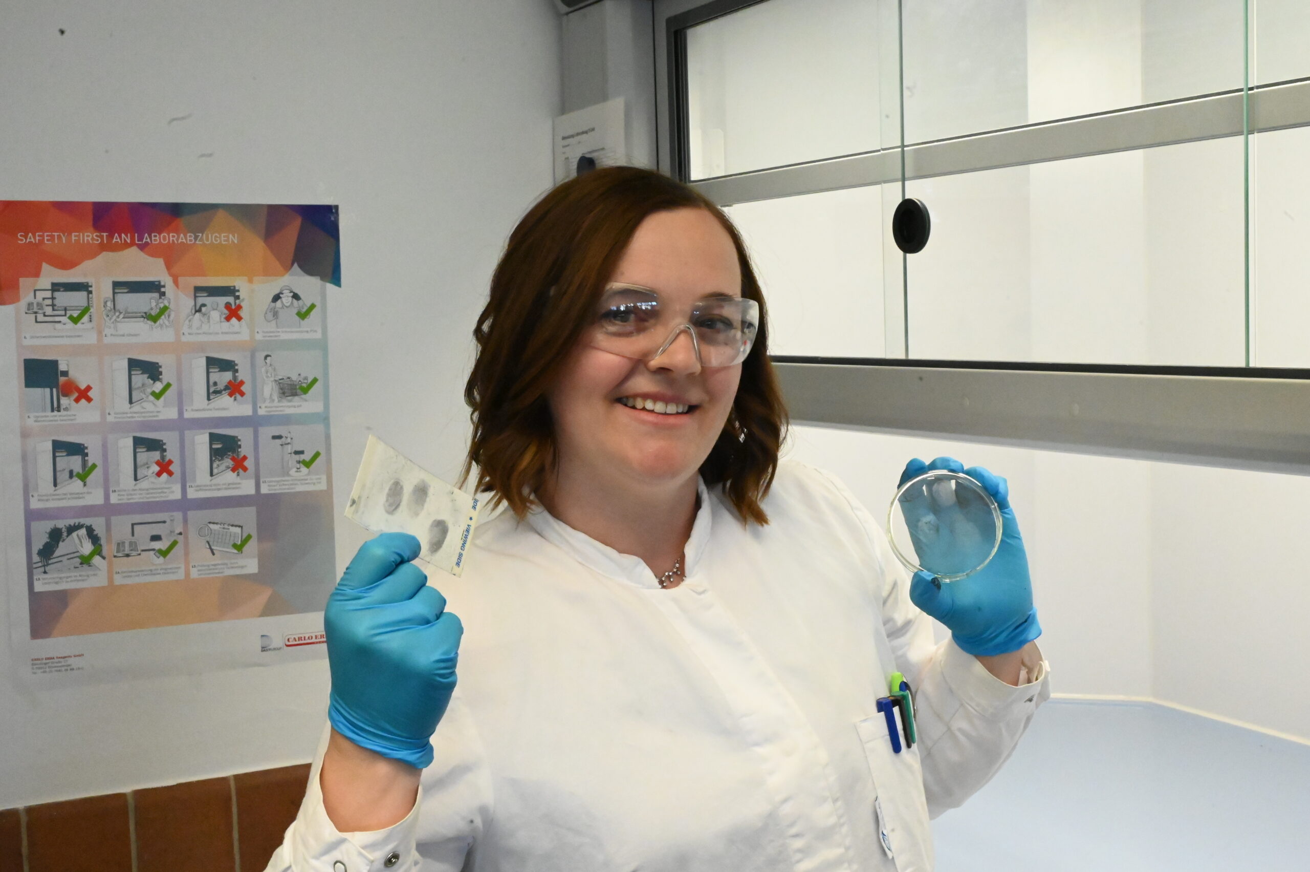 Ein Wissenschaftler mit Schutzbrille, blauen Handschuhen und weißem Laborkittel lächelt, während er eine Petrischale und Klarsichtfolien in der Hand hält. Er steht in einem Labor mit einem TVO-Sicherheitsposter an der Wand. at Hochschule Coburg