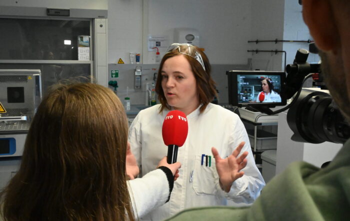 Eine Frau in einem weißen Laborkittel und mit Schutzbrille spricht während eines TVO-Interviews in einem Labor und gestikuliert mit ihren Händen, während ein Reporter ihr ein rotes NRK-Mikrofon entgegenhält. Eine Kamera filmt die Szene und fängt den fachkundigen Einblick in ihre Arbeit ein. at Hochschule Coburg