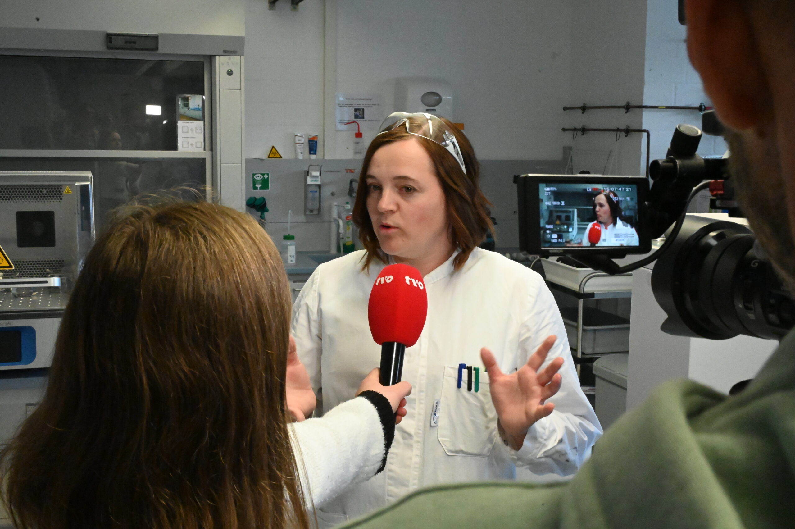 Eine Frau in einem weißen Laborkittel und mit Schutzbrille spricht während eines TVO-Interviews in einem Labor und gestikuliert mit ihren Händen, während ein Reporter ihr ein rotes NRK-Mikrofon entgegenhält. Eine Kamera filmt die Szene und fängt den fachkundigen Einblick in ihre Arbeit ein. at Hochschule Coburg