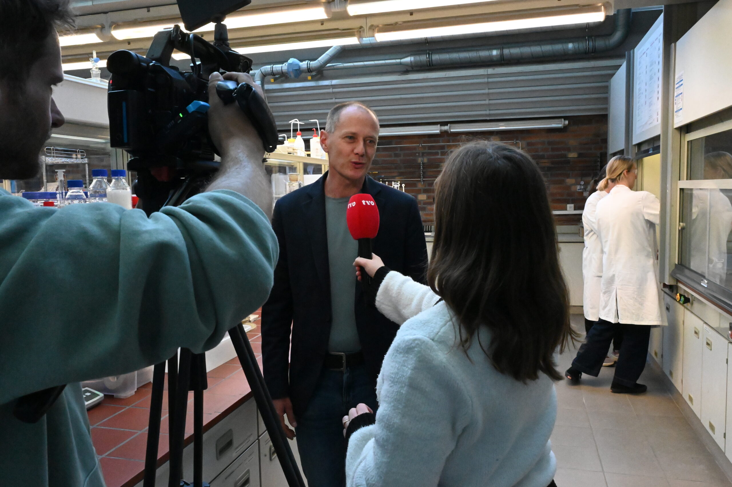 Ein Mann in einem Blazer wird von einer Frau mit einem roten Mikrofon interviewt, möglicherweise für TVO oder LNdW, während ein Kameramann die beiden in einem Labor filmt. Eine weitere Person in einem weißen Laborkittel arbeitet in der Nähe und bietet einen Einblick in das wissenschaftliche Umfeld. at Hochschule Coburg