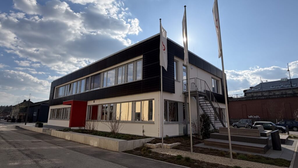 Ein modernes zweistöckiges Gebäude an der Hochschule Coburg hat eine dunkle und weiße Außenverkleidung, große Fenster und eine Außentreppe. Vor dem Gebäude stehen drei weiße Fahnen, während hinter dem Gebäude das Sonnenlicht durch den teilweise bewölkten Himmel bricht. at Hochschule Coburg